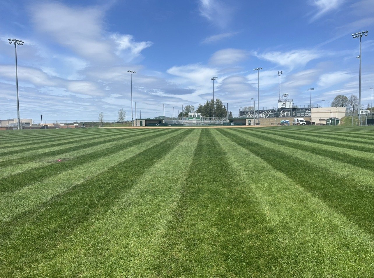A baseball field with freshly mowed grass in striped patterns managed by Ornell lawn services. , under a partly cloudy blue sky, with stadium lights and bleachers in the background.
