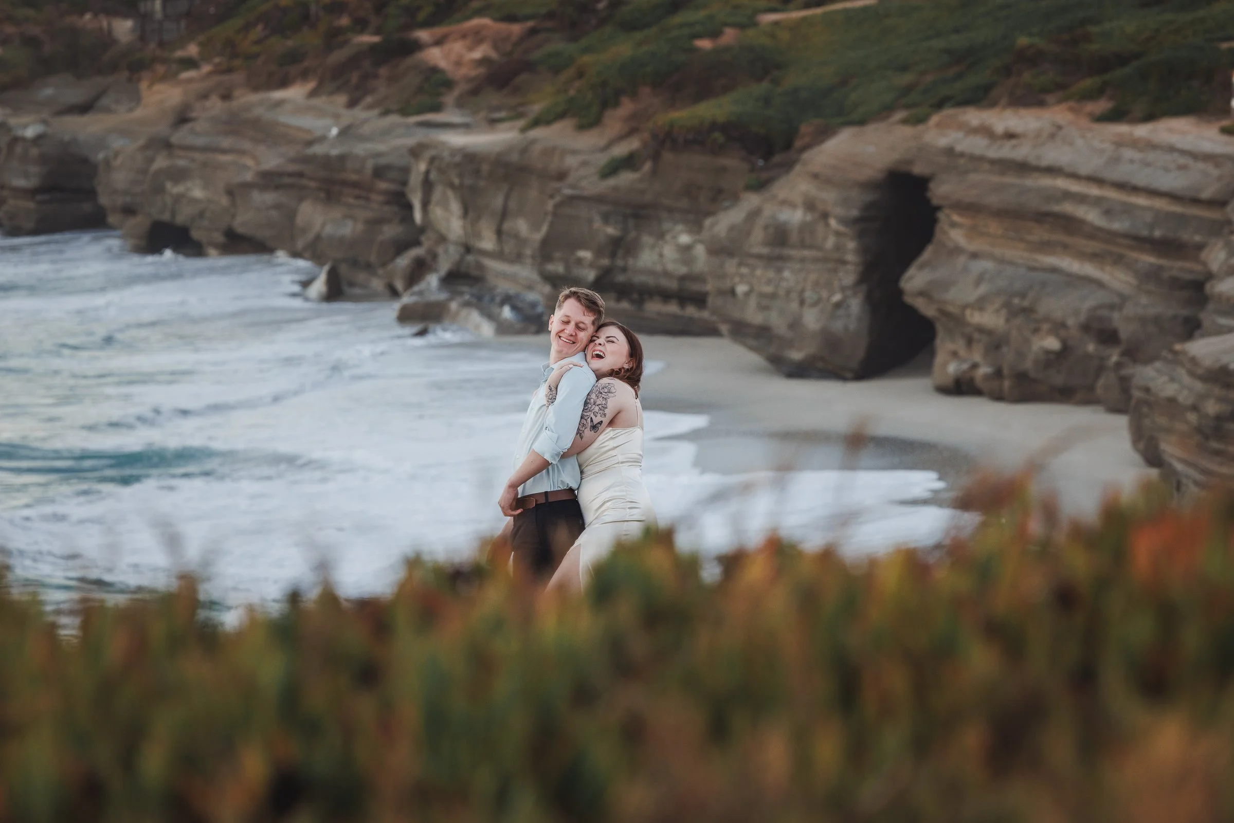 A couple hugging and smiling on a beach with rocky cliffs in the background.