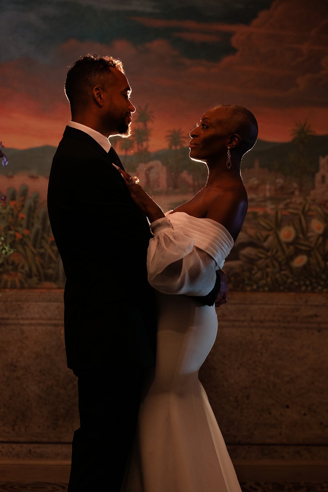 A couple dressed in wedding attire looking at each other closely during a sunset or evening event with a scenic backdrop.