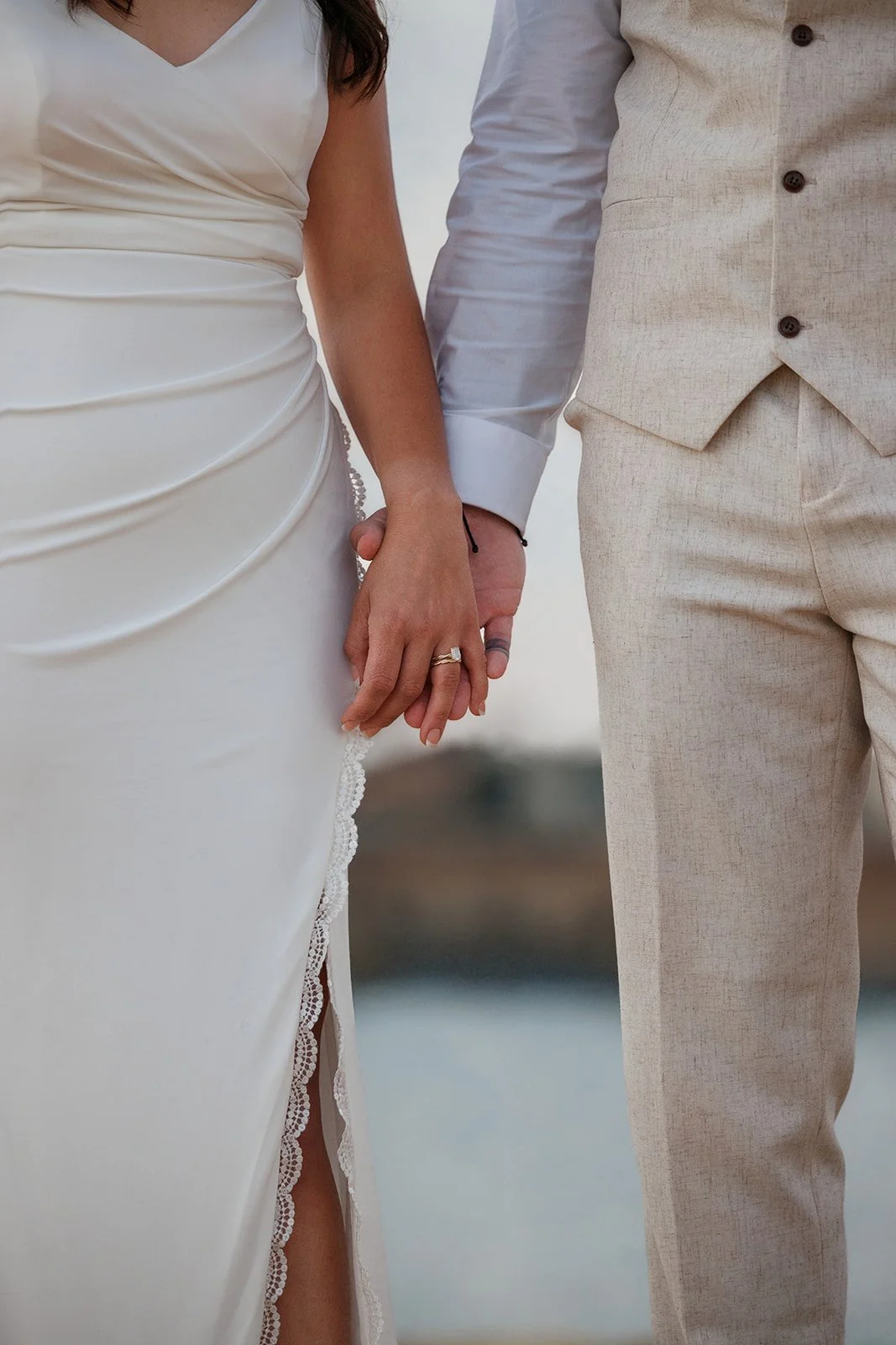 Close-up of a bride and groom holding hands, showing wedding rings. The bride wears a white satin dress with lace trim, and the groom wears a beige linen suit.