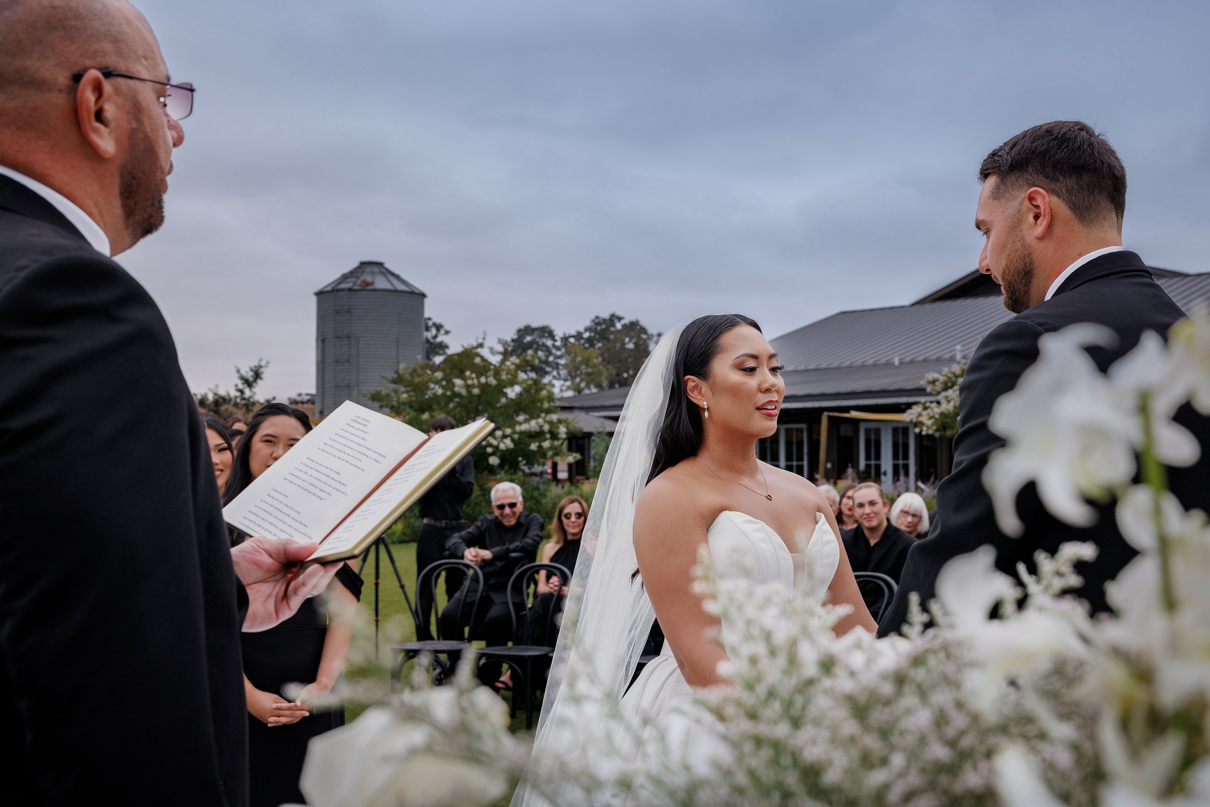 A bride and groom exchange vows during an outdoor wedding ceremony, with guests sitting on chairs behind them and a man reading from a book on the left, under a cloudy sky.