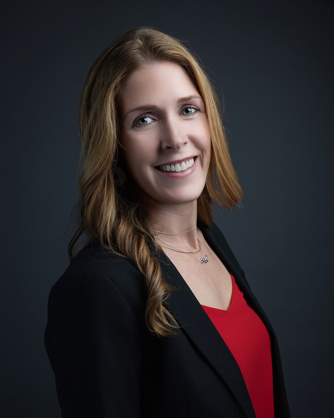 Portrait of a woman with reddish-brown hair, smiling, wearing a black blazer and a red top, against a dark background.