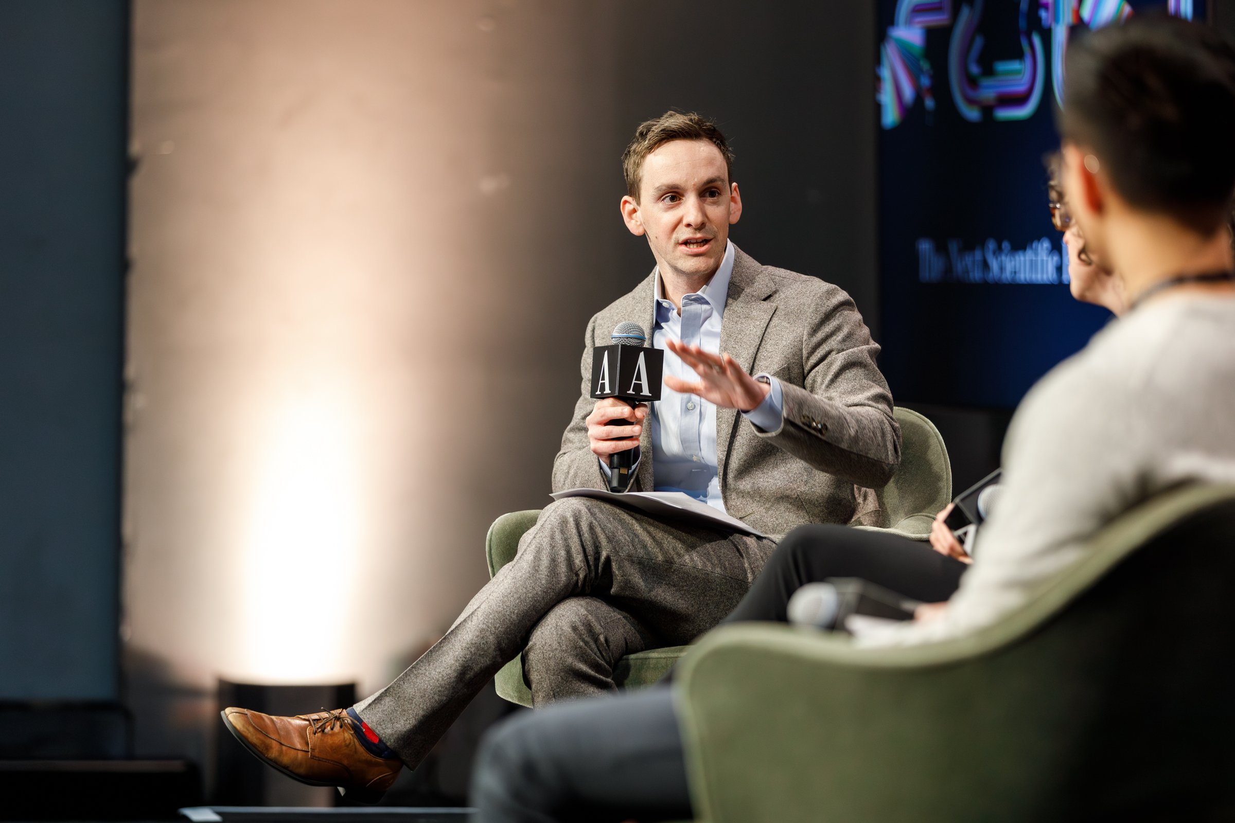A man in a gray suit holding a microphone and talking during a panel discussion at an event, with other participants in the background.
