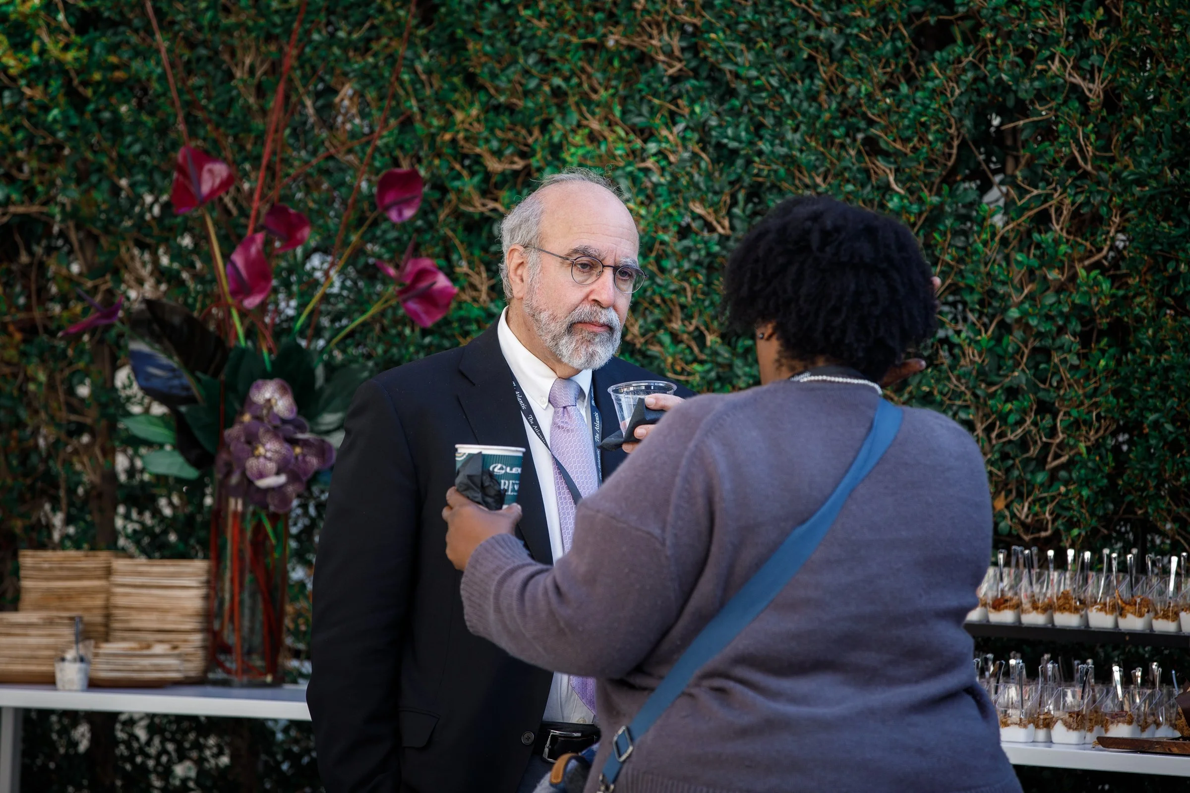A man in a suit and glasses is having a conversation with a woman with curly hair at an outdoor event, with a background of greenery and a table with drinks and snacks.