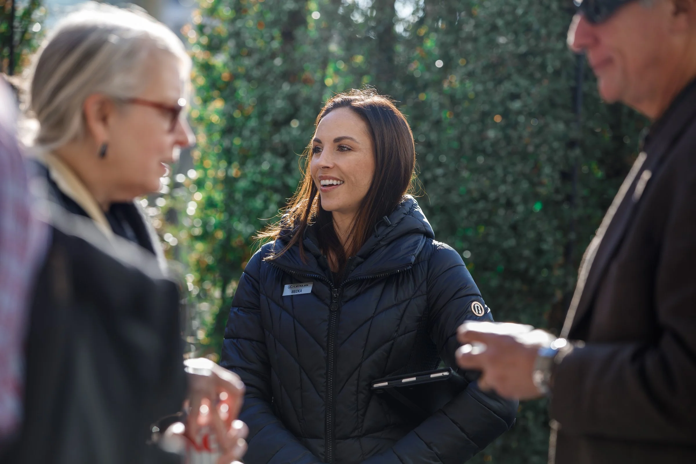 A woman in a blue jacket smiling and talking to a man in sunglasses and a dark jacket while outdoors.