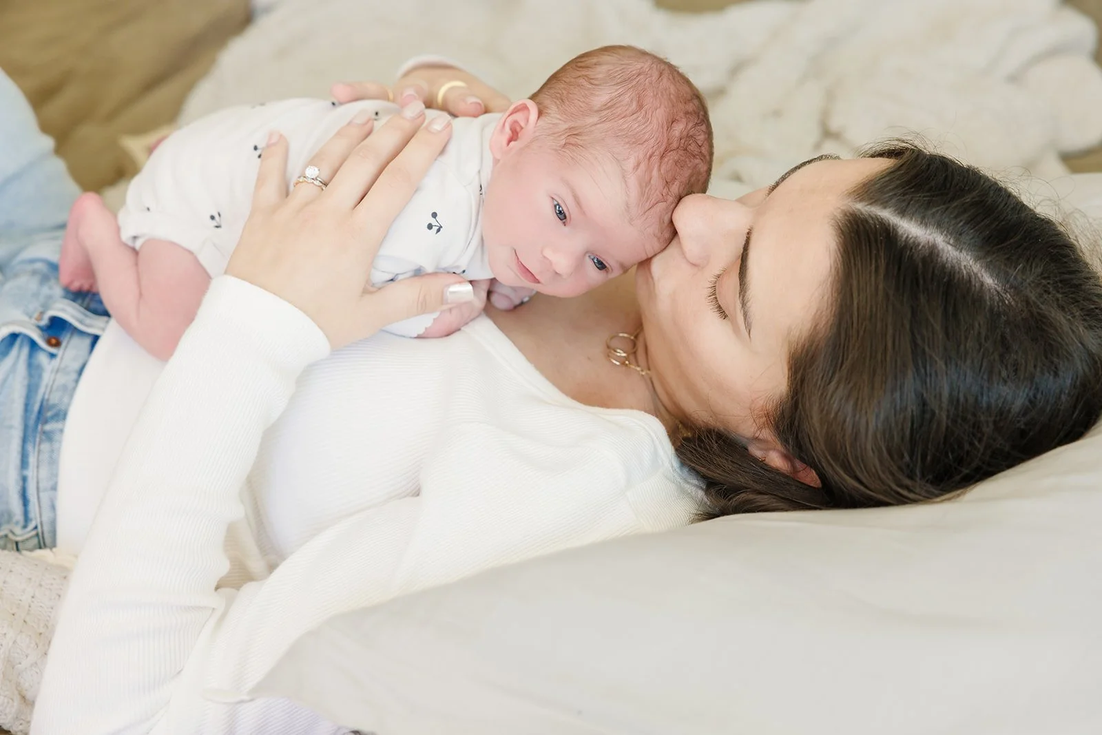 A woman with dark hair lying on her side, holding a baby close to her face and kissing the baby's forehead.