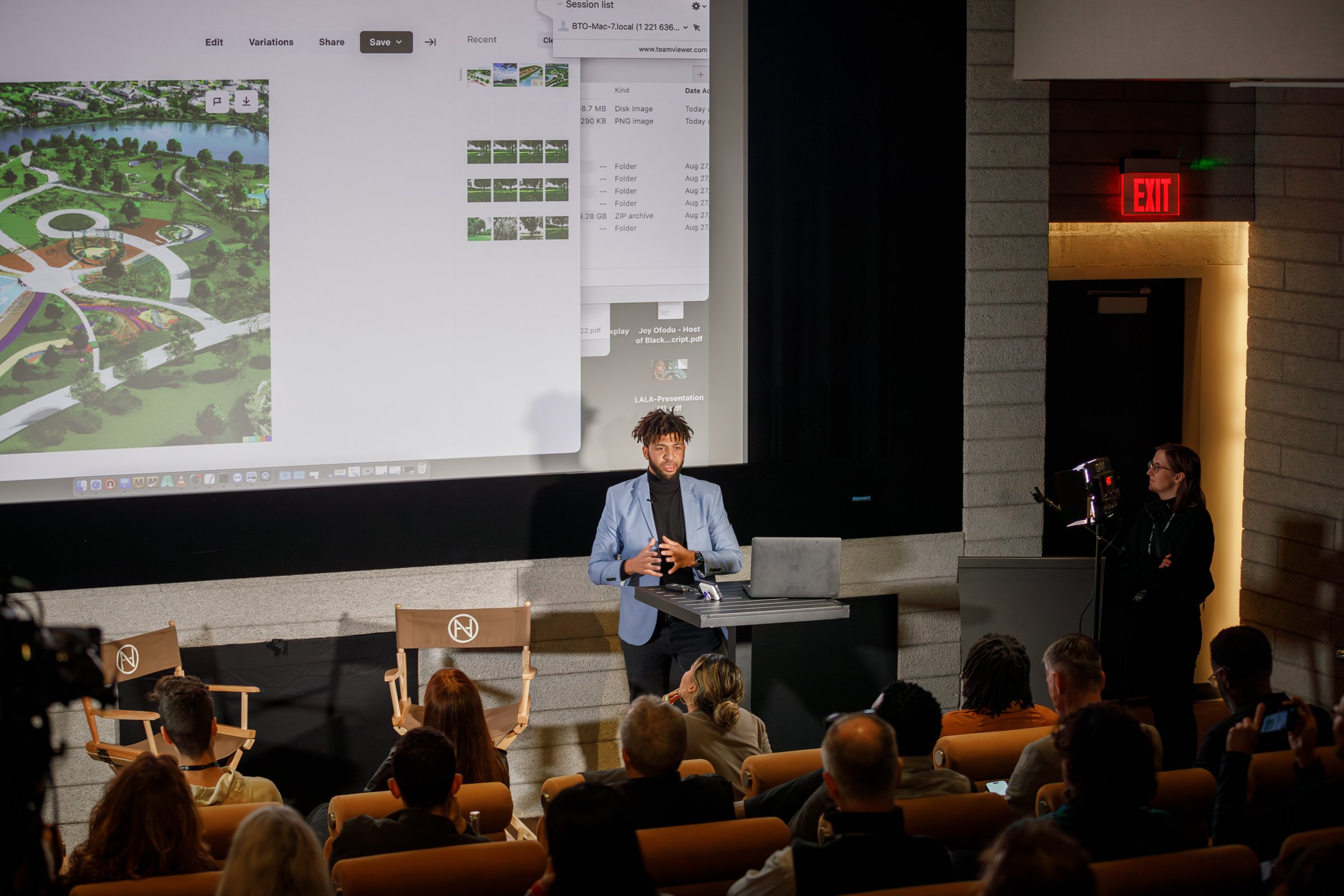 A man in a blue blazer giving a presentation in front of an audience in a theater. Behind him is a large screen displaying a map or diagram. An woman stands near a light panel on the right. The audience is seated and watching attentively. There is an
