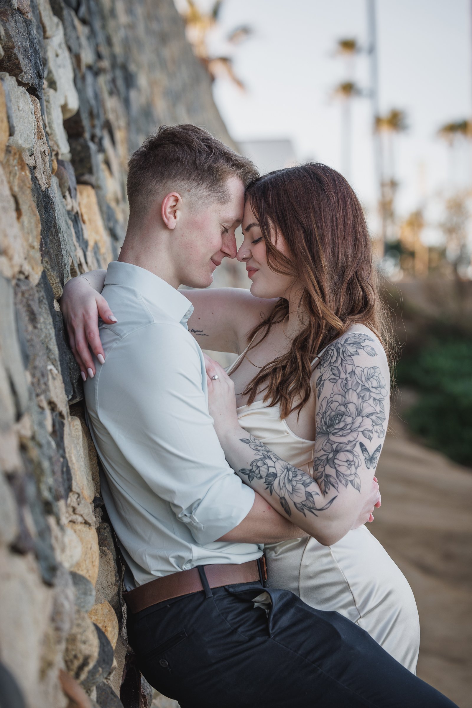 A couple with closed eyes and gentle smiles, standing close with foreheads touching, outdoors next to a stone wall, the woman with floral tattoos on her arms, appears happy and intimate.