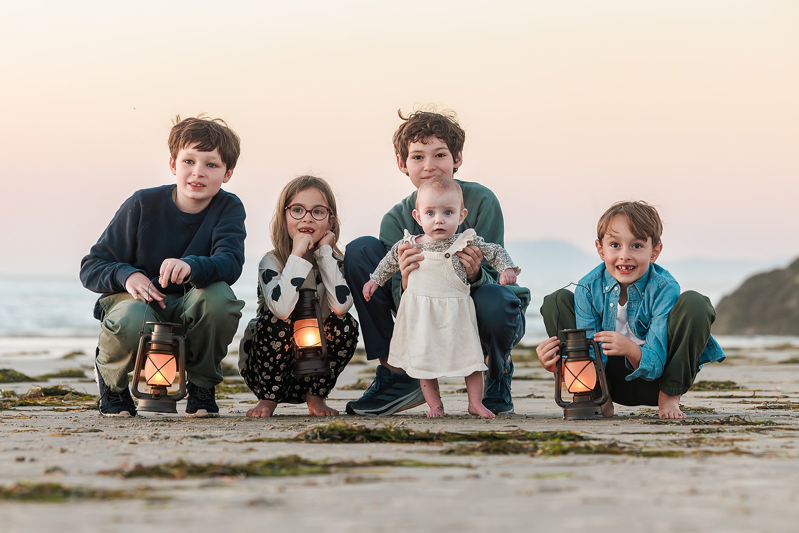 Group of five children, two boys, two girls, and a baby girl, sitting and kneeling on a beach at dusk, holding lanterns with glowing lights, with a rocky shoreline and ocean in the background.
