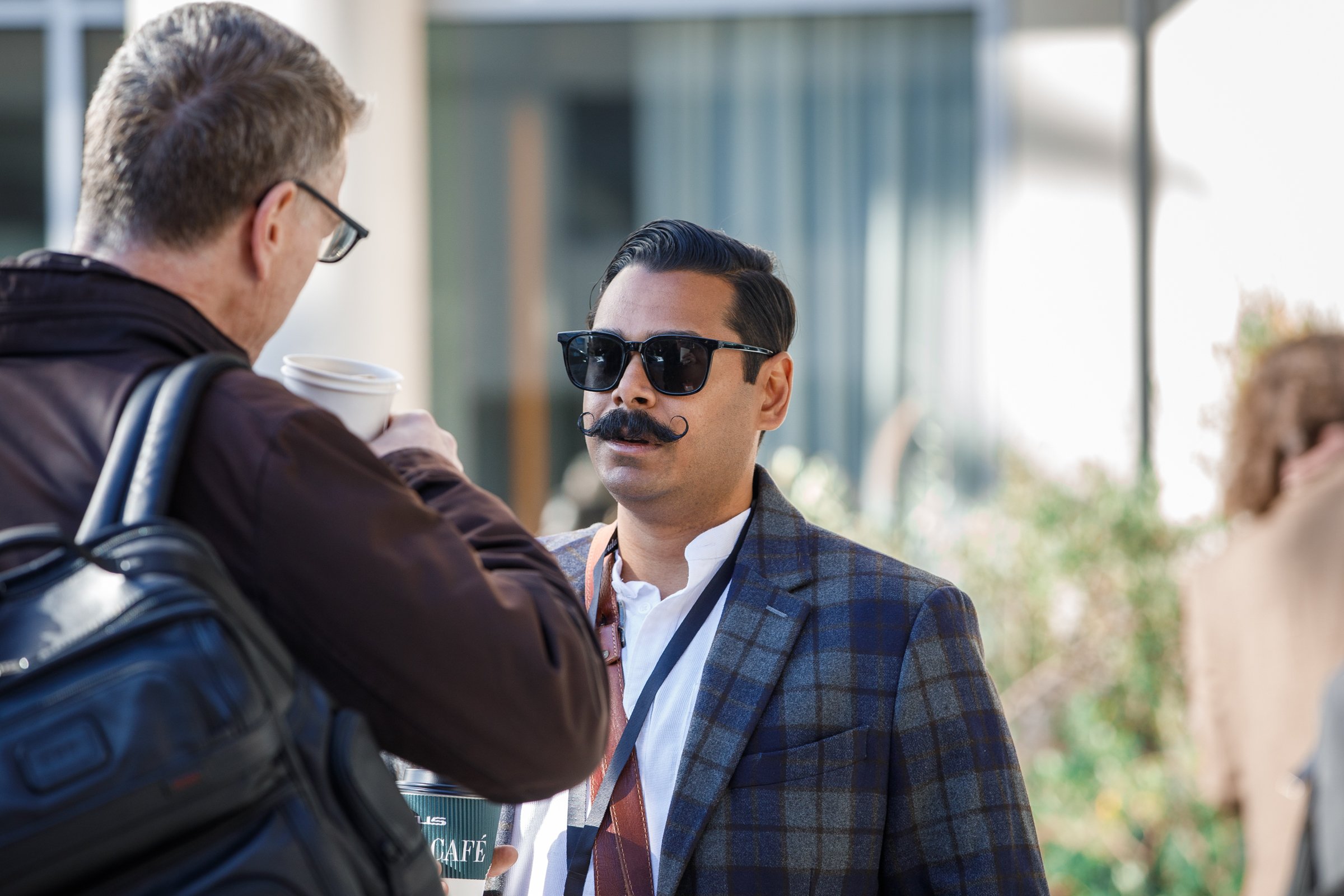 Man with dark hair, sunglasses, and a mustache, wearing a plaid blazer, being approached by another man holding coffee cups.