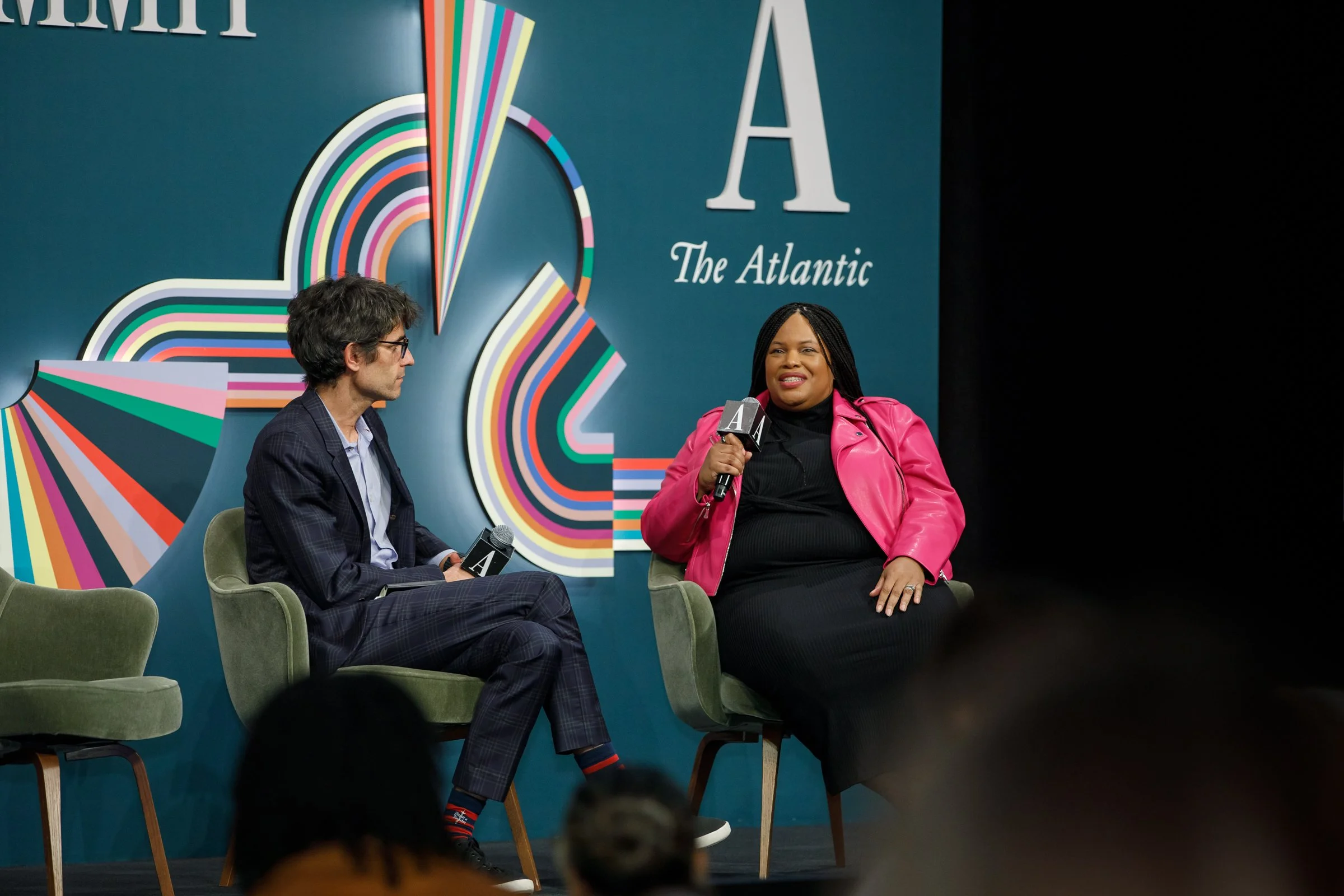 Two individuals seated on stage in discussion at an event hosted by The Atlantic. The woman on the right, wearing a pink jacket, is speaking into a microphone. The man on the left, dressed in a dark suit, listens attentively. A colorful, abstract gra