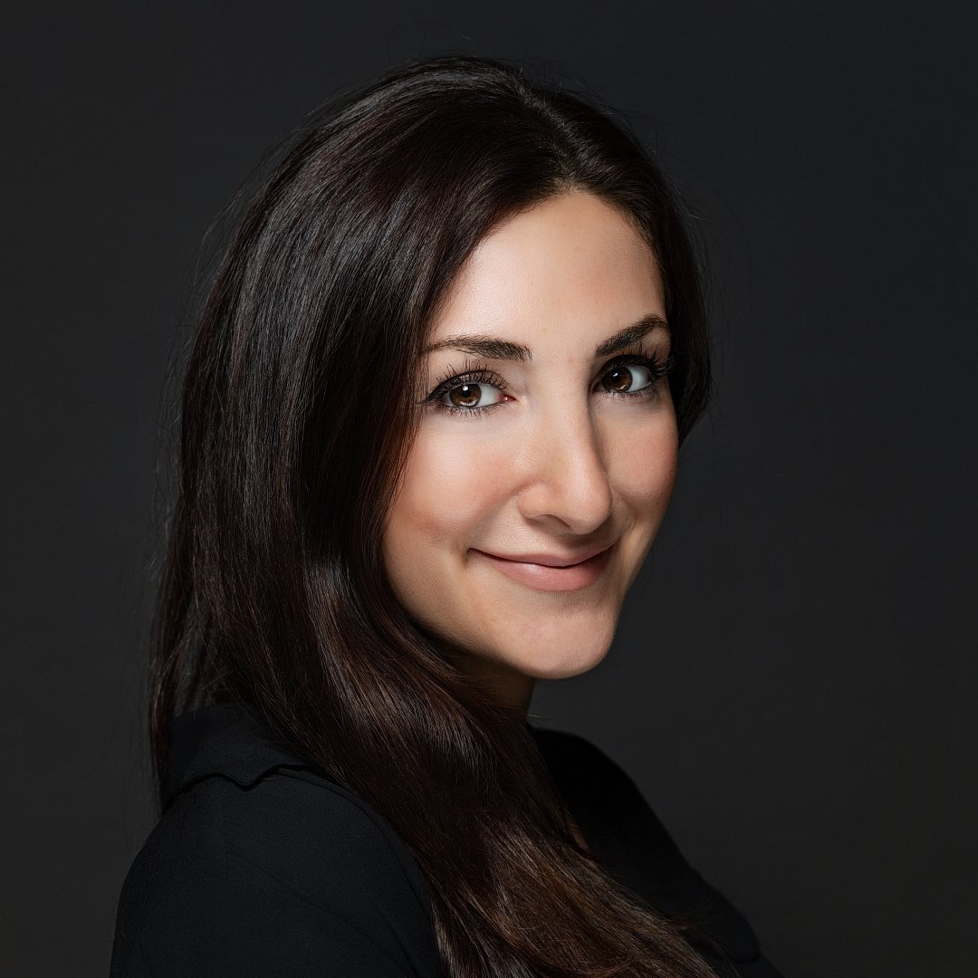 Close-up portrait of a woman with long, dark hair and a slight smile, against a dark background.