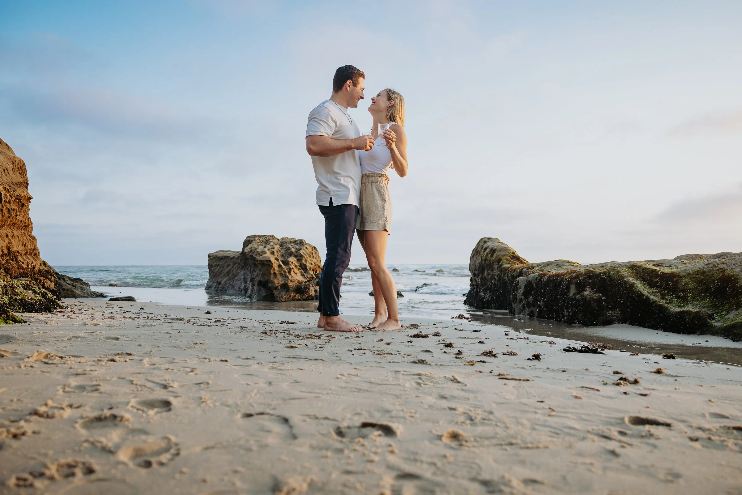 A couple standing barefoot on the beach near rocks, smiling and looking at each other