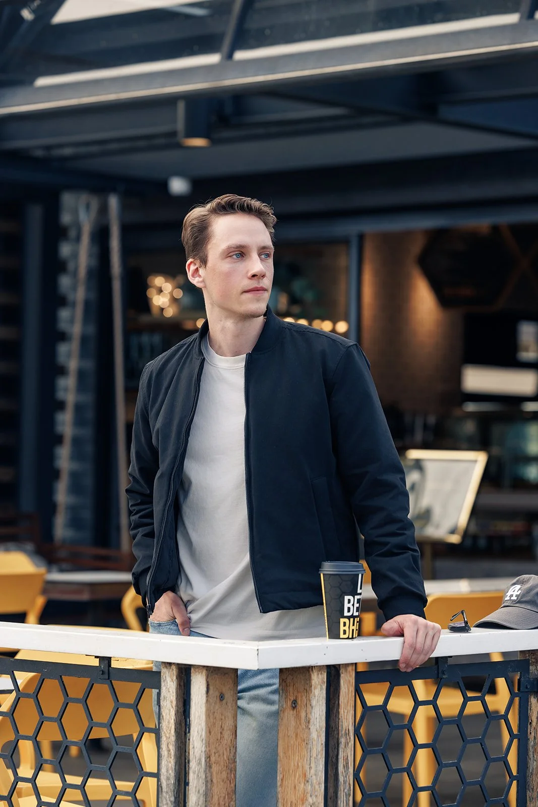 A young man with light skin, short brown hair, and blue eyes stands outdoors, leaning on a railing with one hand. He is wearing a black jacket over a white T-shirt. On the railing next to him, there is a black disposable coffee cup with yellow and wh