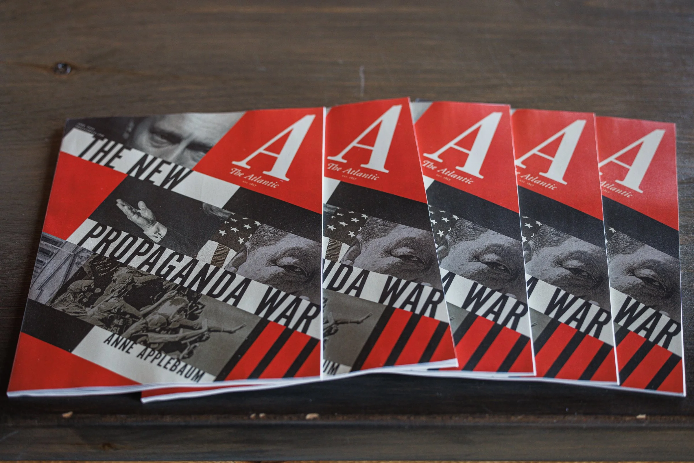 A stack of magazines titled 'The Atlantic' with a cover story about propaganda war featuring a black-and-white photo of a man covering one eye, a waving American flag, and an image of soldiers in combat, on a dark wooden surface.