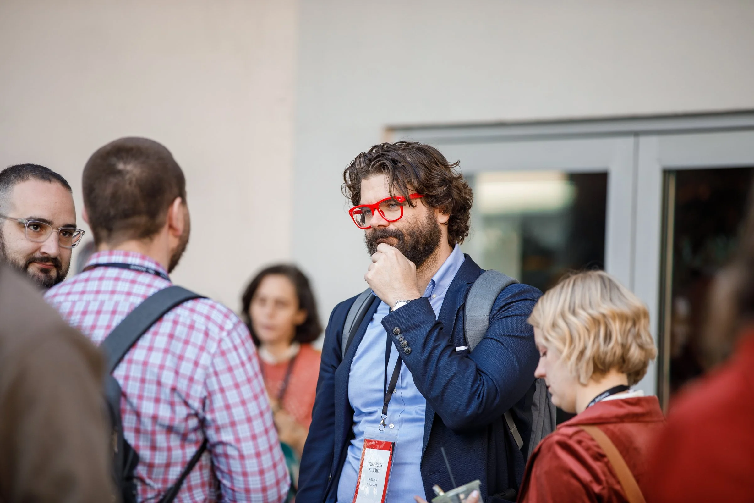 A group of people at an indoor event, including a man with curly hair, beard, and red glasses who is thinking with his hand on his chin.