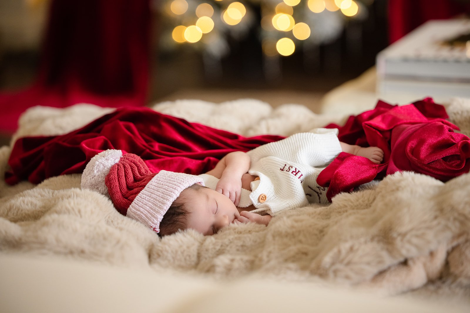 A sleeping baby wearing a Santa hat and a white sweater, lying on a fluffy blanket surrounded by Christmas decorations, including a red velvet cloth, with blurred Christmas lights in the background.