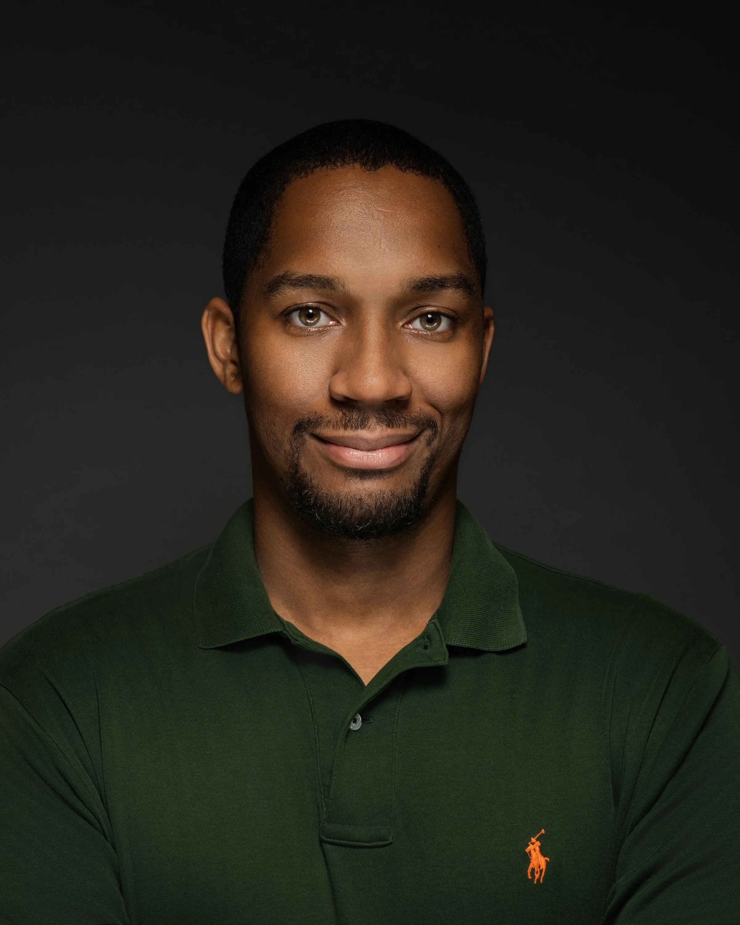 Portrait of a smiling man wearing a dark green polo shirt with a small orange logo, against a dark background.