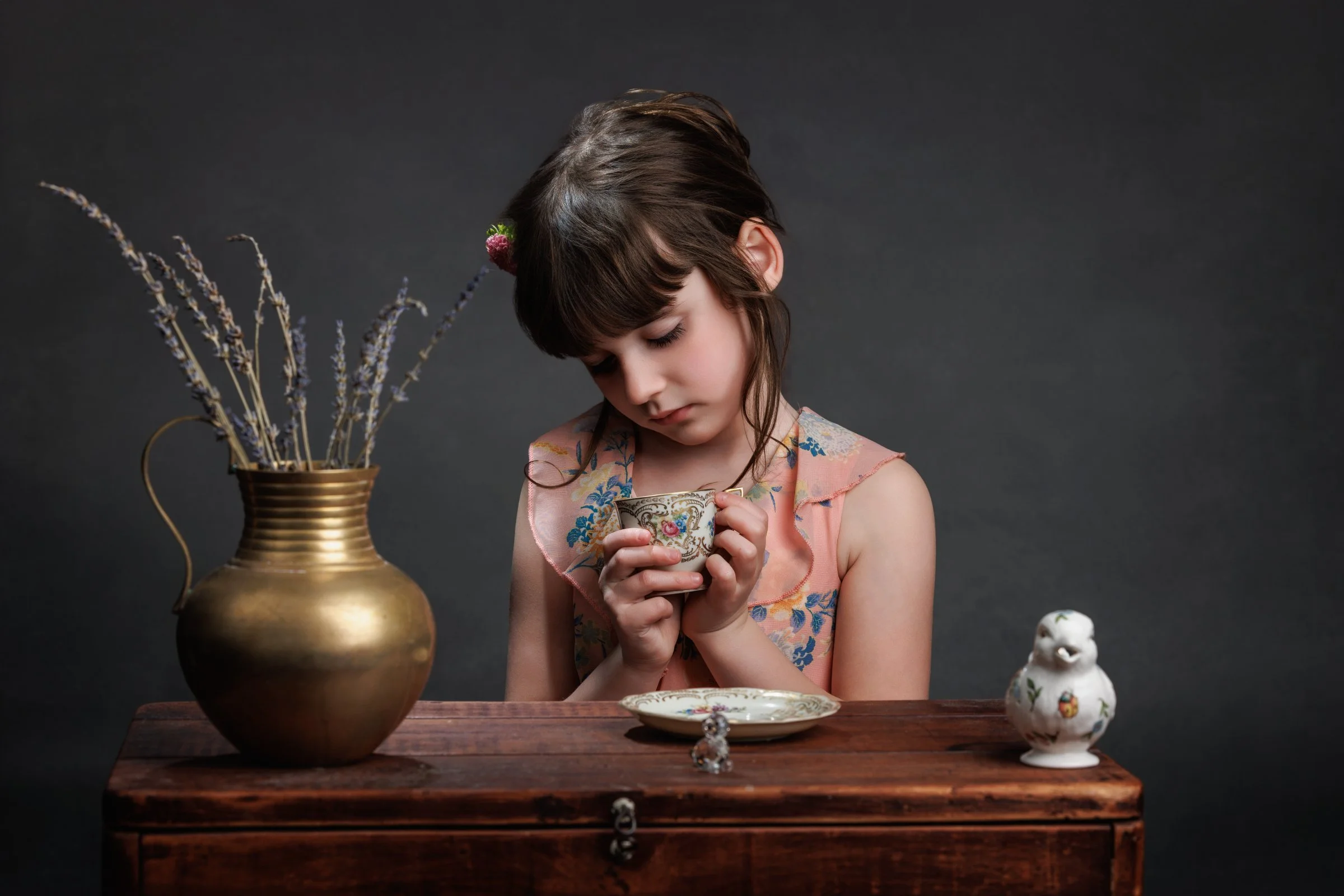 A young girl with dark hair and bangs wearing a pink floral dress, holding a decorative teacup, sitting at a wooden table with a plate, a brass vase with lavender, and a ceramic bird figurine, against a dark background.