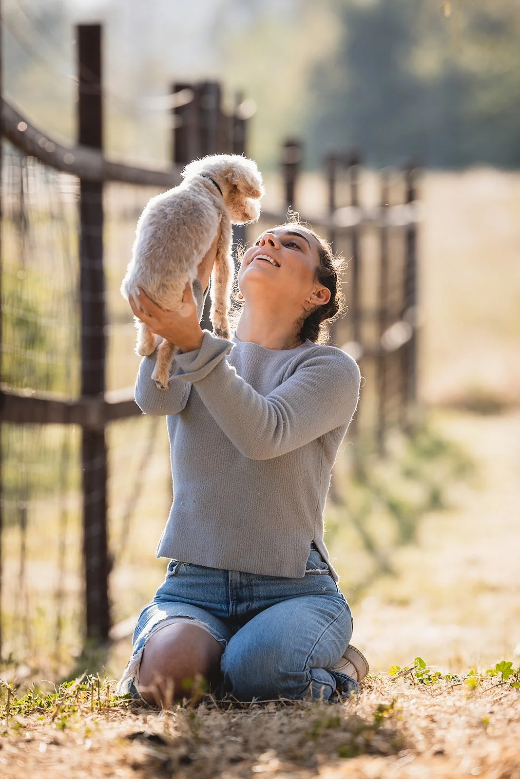A woman kneeling on the ground, holding a small puppy up in the air, outdoors on a sunny day.