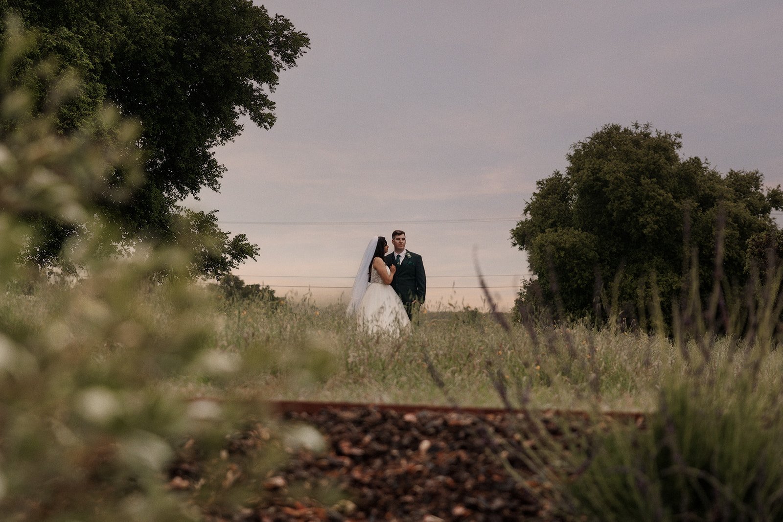 A bride and groom stand together in a field surrounded by trees, with a cloudy sky overhead, during their wedding photo shoot.