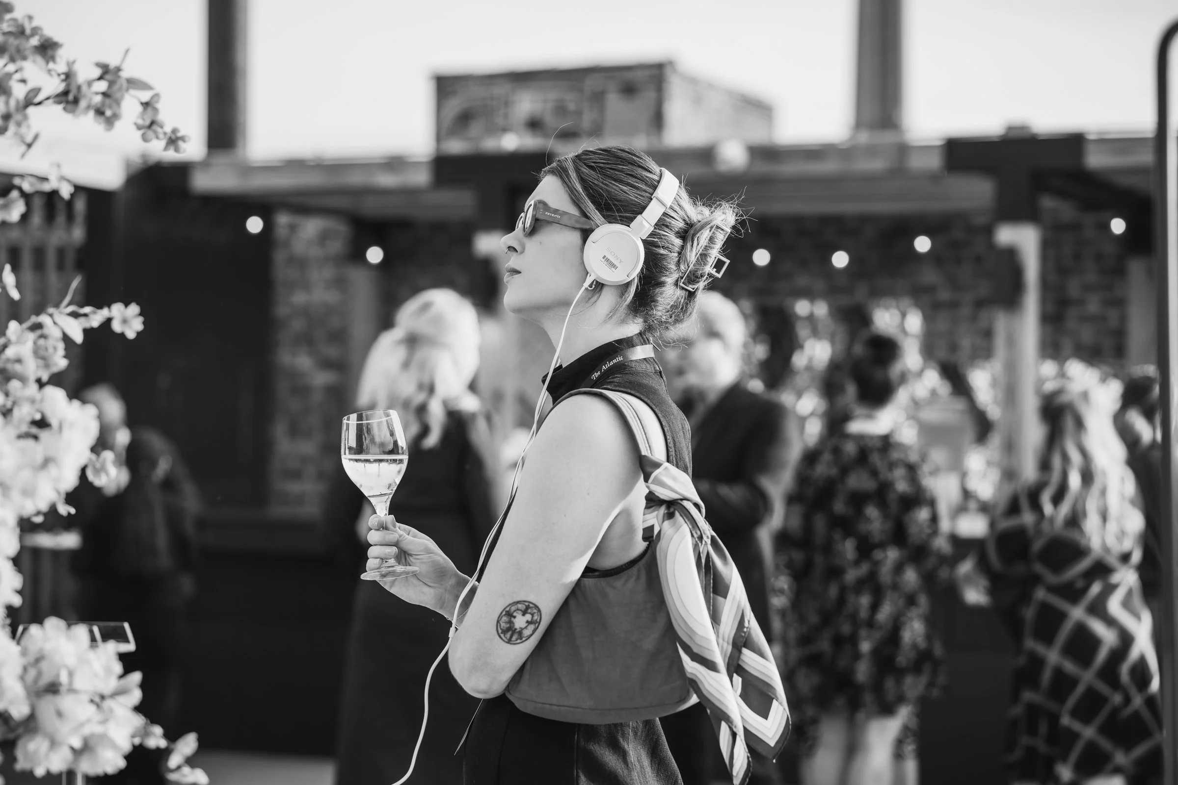 A woman with sunglasses and a tattoo on her arm, listening to music with headphones, holding a glass of white wine at a social gathering on an outdoor terrace.