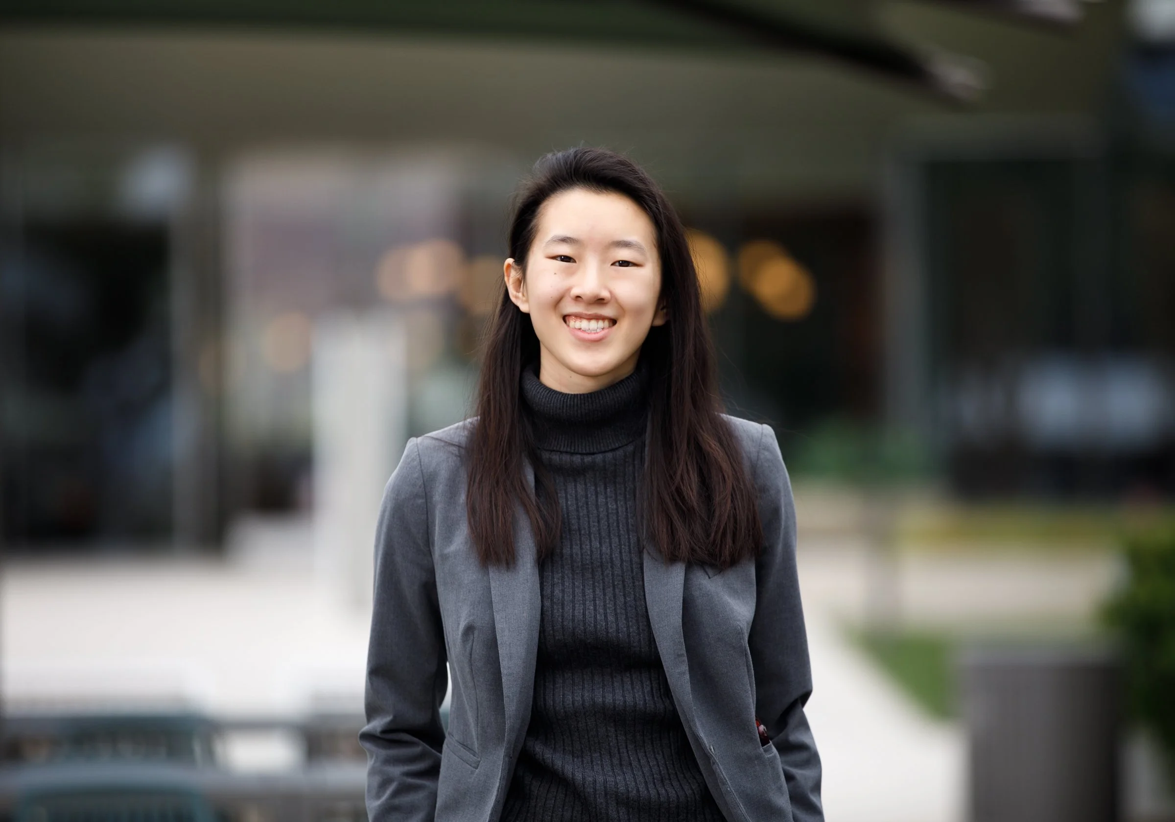 Young Asian woman with long dark hair smiling outdoors, wearing a gray blazer and black turtleneck, with a blurred urban background.