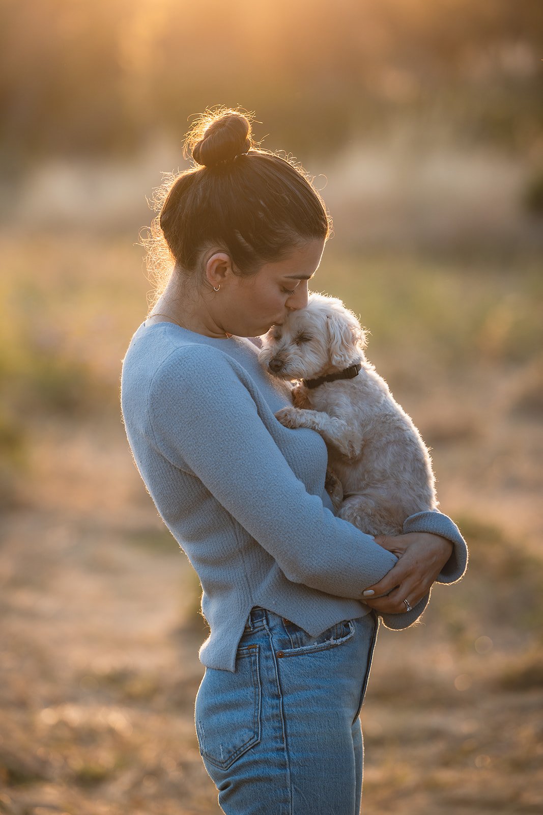 A woman in a blue sweater holding a small white puppy during sunset in an outdoor setting.