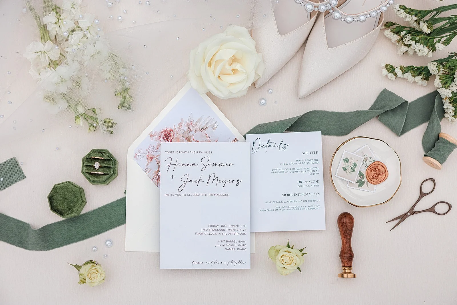 Wedding invitation and accessories on a table, including a white rose, pearl necklace, white shoes, green ribbon, postage stamps, scissors, and a wax seal stamp.