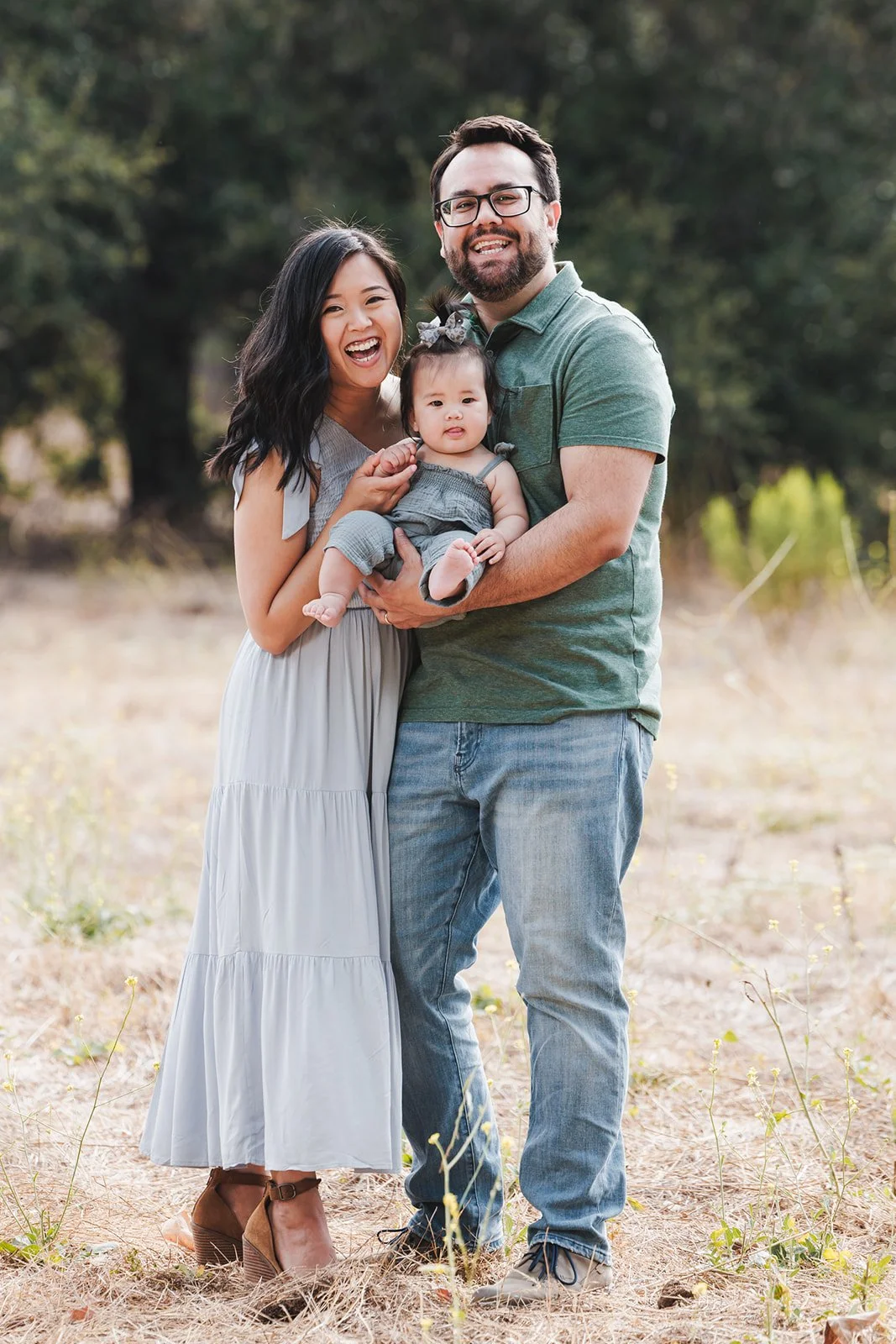 Happy family of three standing outdoors, with tall trees in the background. The woman has long dark hair and is smiling widely, wearing a light gray maxi dress and brown high heels. The man has dark hair, a beard, and glasses, wearing a green polo sh
