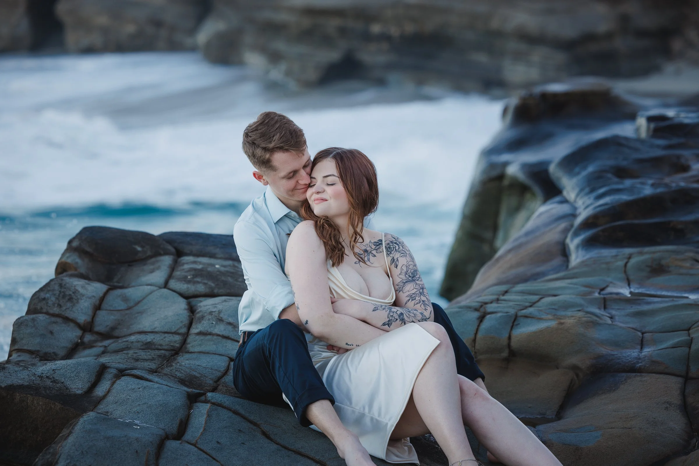 A couple sitting on rocks by the ocean with the man behind the woman, both with closed eyes, smiling softly.