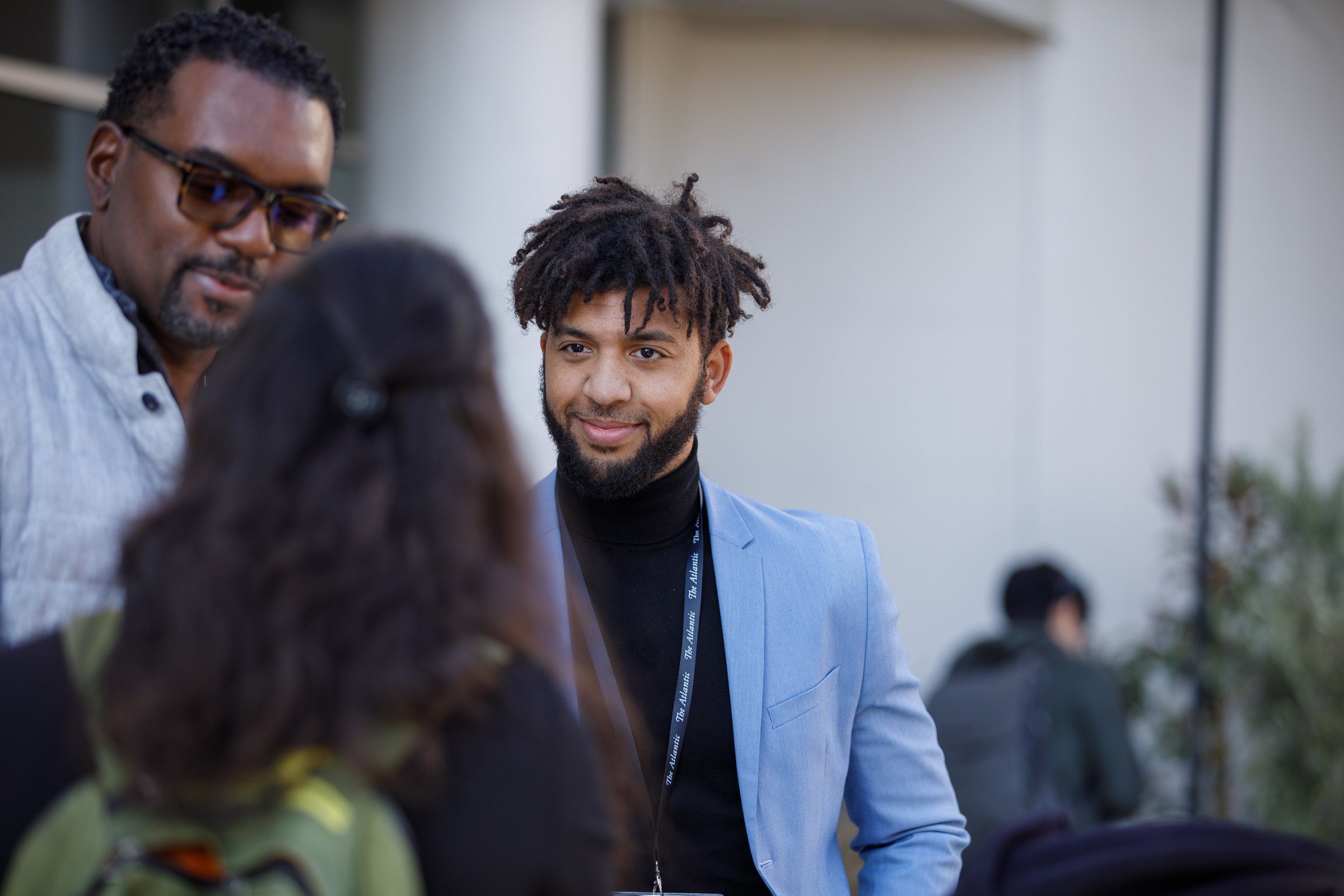 Three people having a conversation at a professional event. A man in a light blue blazer with a black turtleneck and a lanyard is smiling, and another man with glasses and a gray shirt is engaged with the woman whose back is to the camera, with long 