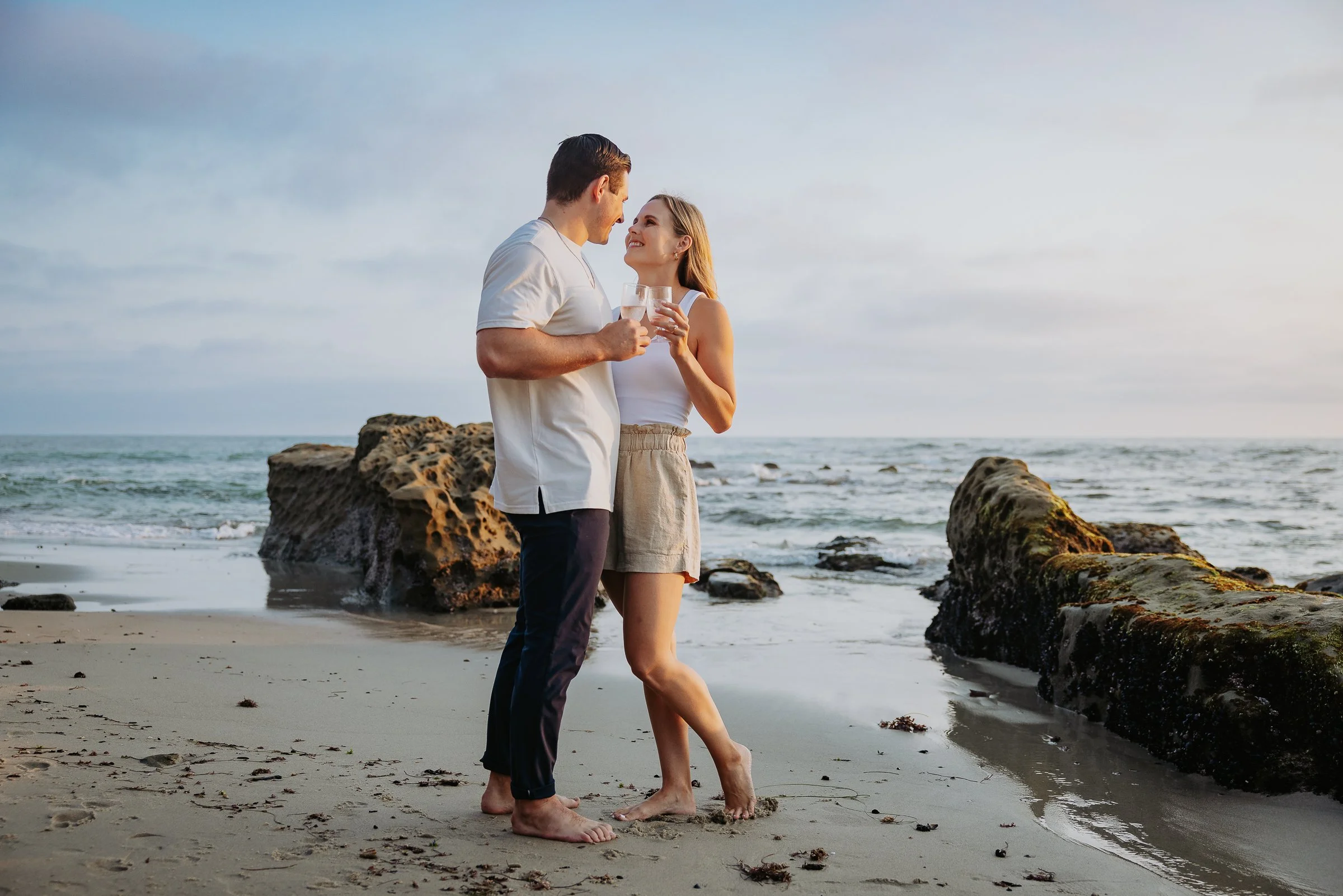 A couple standing barefoot on the beach holding glasses of wine, gazing at each other with the ocean, rocks, and sky in the background during sunset.