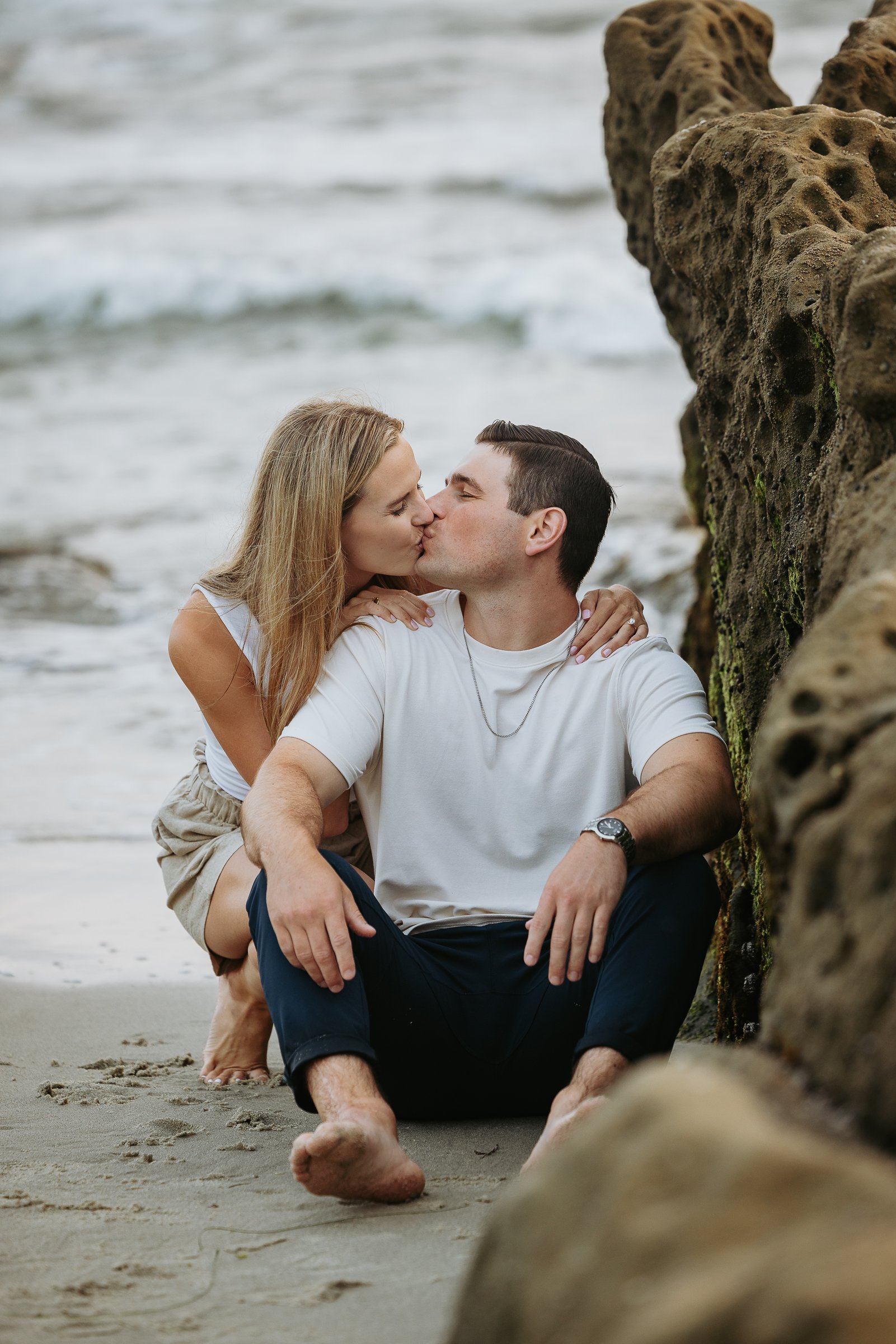 A couple sitting on the beach, kissing, with rocks and ocean waves in the background.