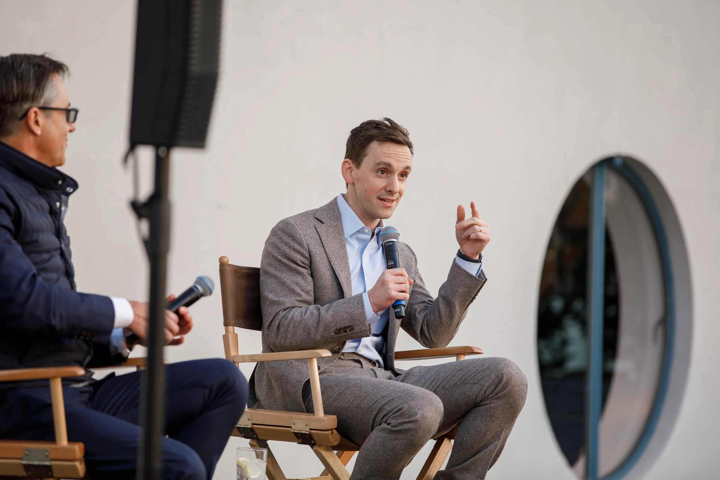 Two men sitting in chairs, participating in a discussion. The man on the right, wearing a light gray suit, is speaking into a microphone and gesturing with his hand. The man on the left, in dark clothing and glasses, is holding a microphone. They are