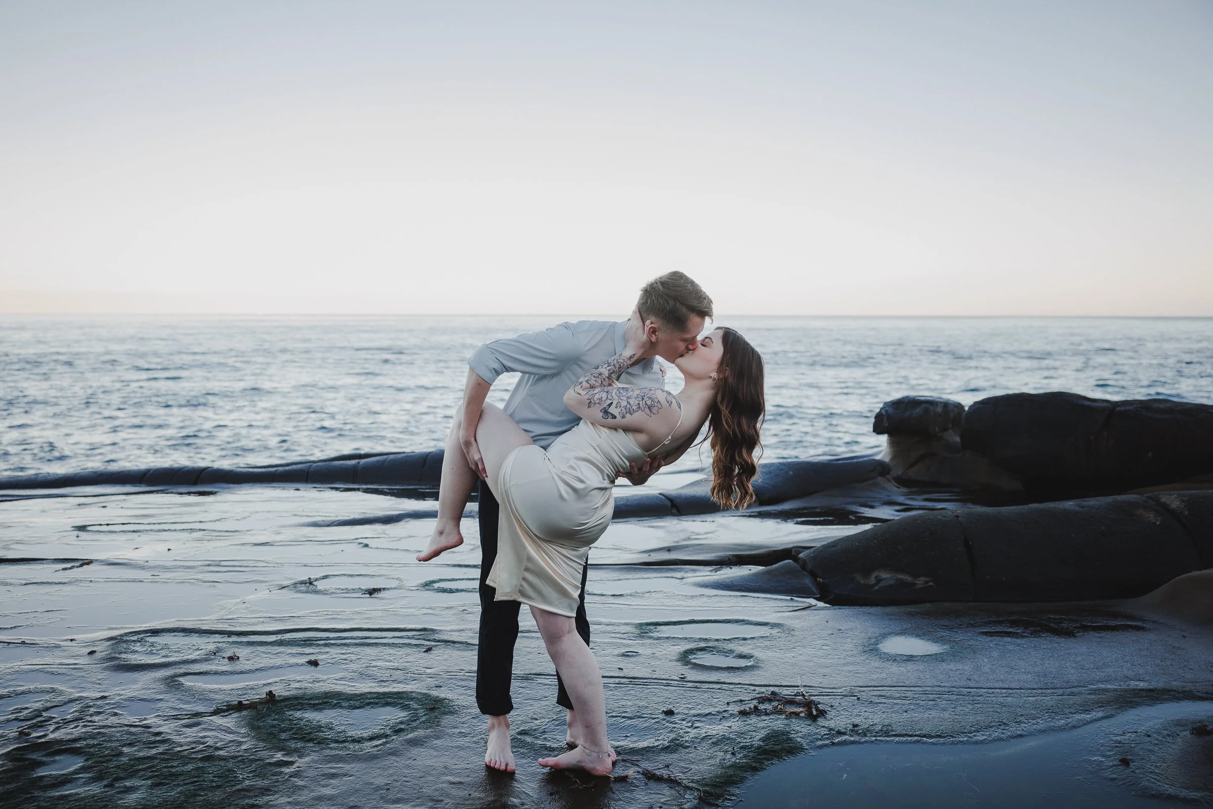 A couple sharing a kiss on a rocky beach at sunset, with the man dipping the woman while standing barefoot in the shallow water.