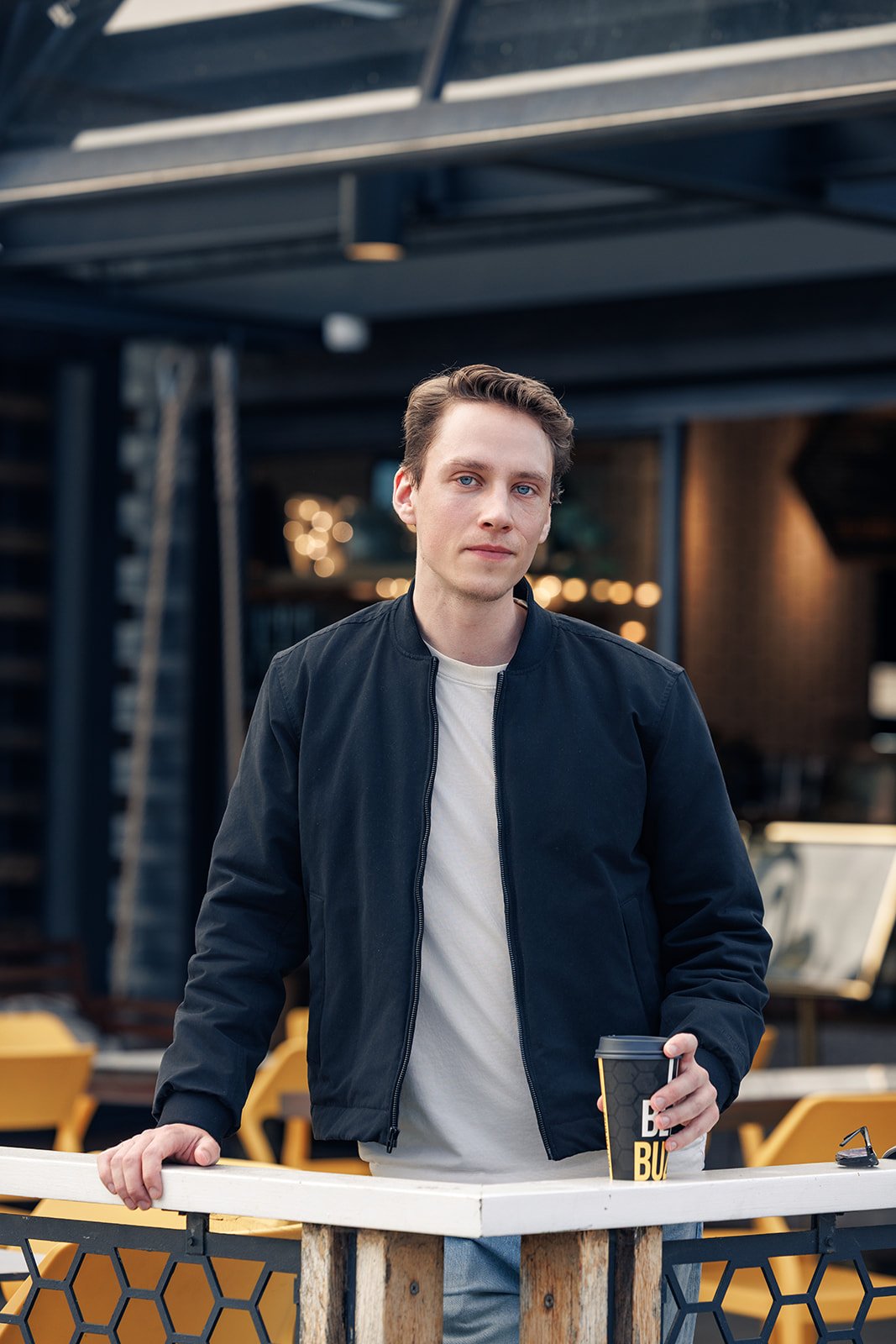 Young man with brown hair and blue eyes wearing a black jacket and white t-shirt, holding a coffee cup, standing outdoors near a white barrier with yellow chairs in the background.