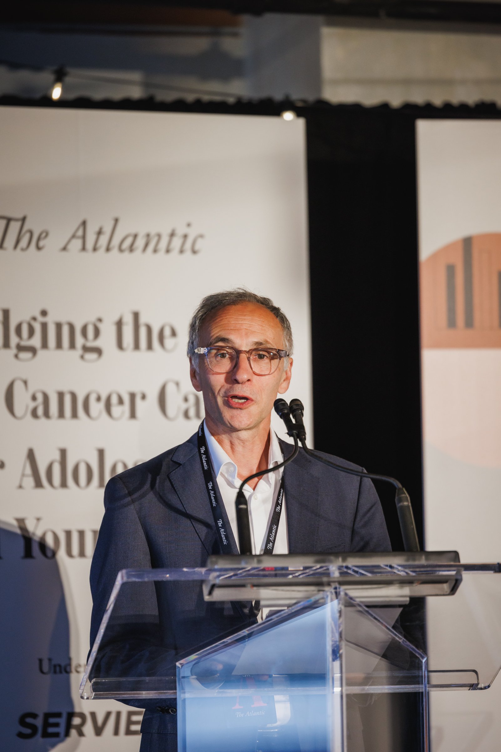 A man in a suit and glasses speaking at a podium at a conference, with banners in the background displaying text related to cancer care and adolescence.