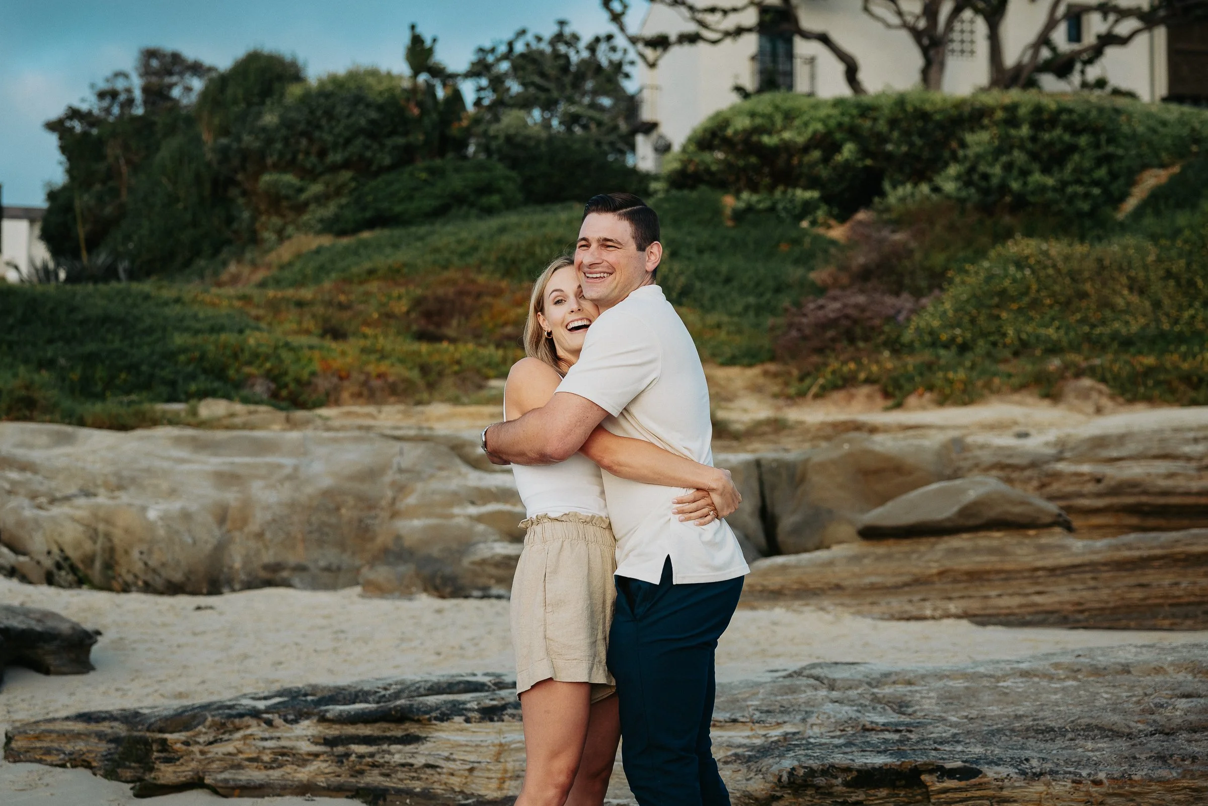 A young couple hugging and smiling on a beach with rocks and greenery in the background.