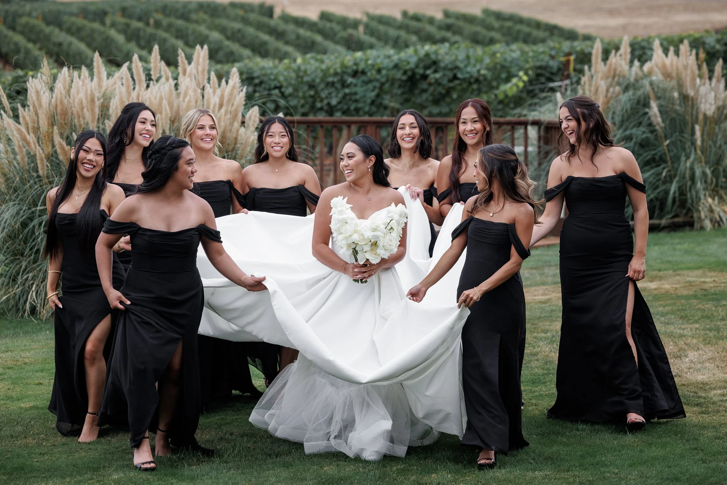 A bride in a white wedding dress holding a bouquet of white flowers, surrounded by ten women in black dresses outdoors on green grass, smiling and laughing.