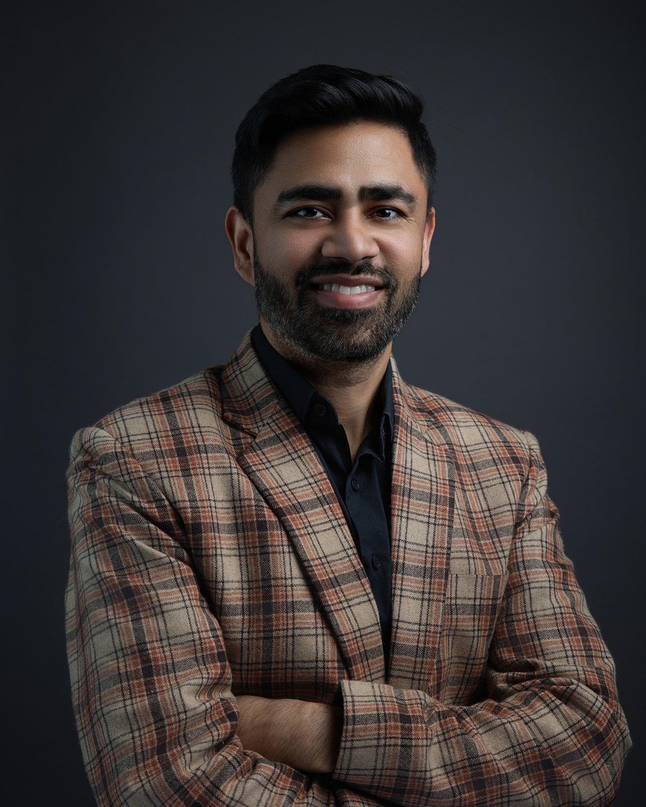 Portrait of a man with dark hair, beard, wearing a checked blazer and black shirt, smiling against a dark background.