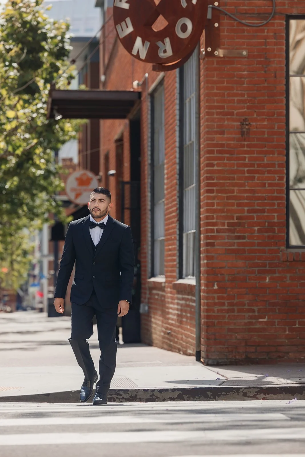 A man in a black tuxedo and bowtie walking across a crosswalk on a city street during daytime.