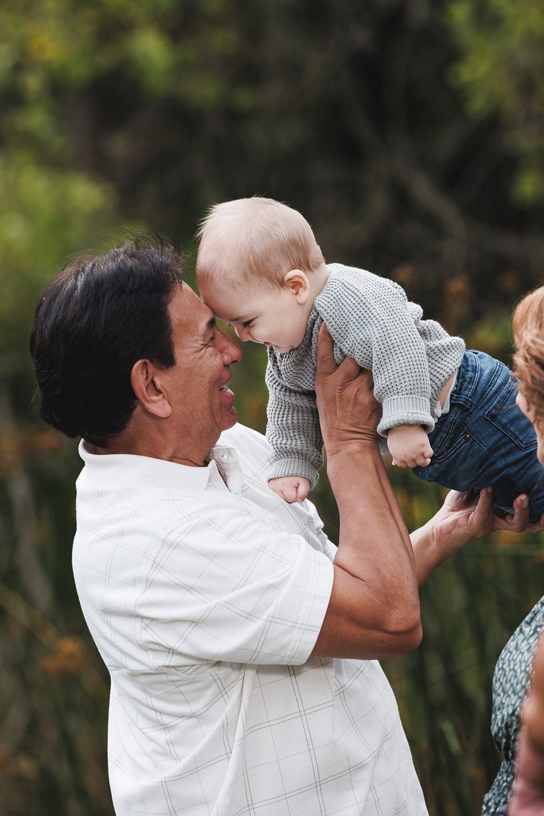 An elderly man with dark hair is smiling and holding a young boy, who is leaning in with their foreheads touching. The boy has blonde hair and is dressed in a gray sweater and blue jeans. The background shows a blurry outdoor setting with green trees
