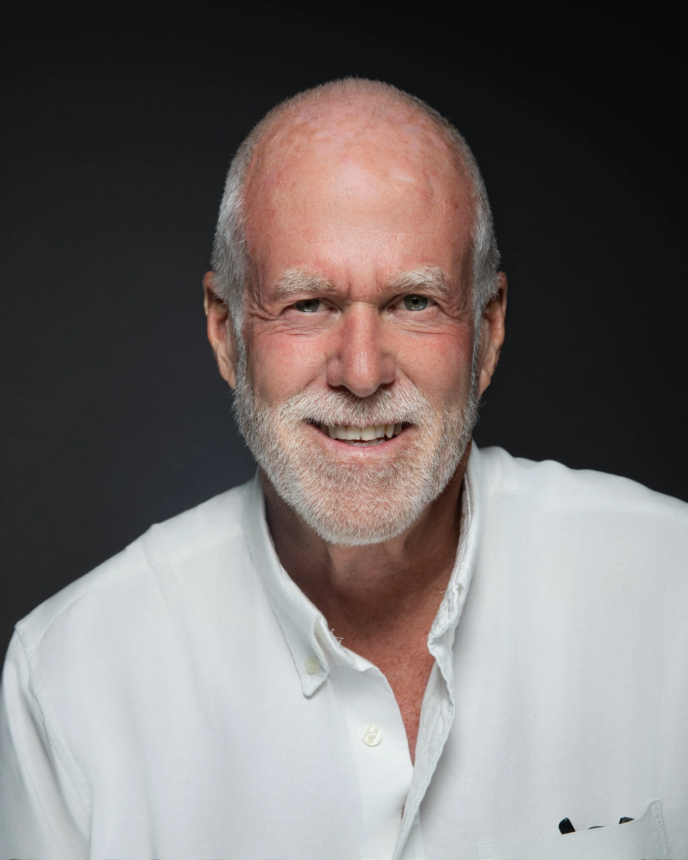 Portrait of an older man with white hair and a beard, wearing a white button-up shirt, smiling against a dark background.