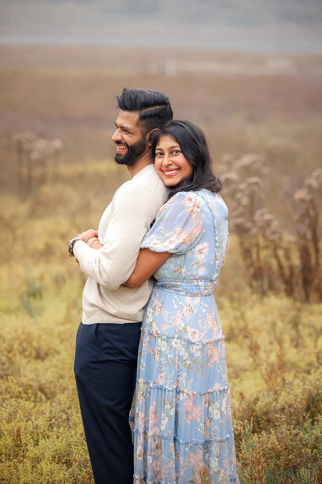 A smiling woman in a blue floral dress hugging a man from behind outdoors, with a blurred grassy field background.