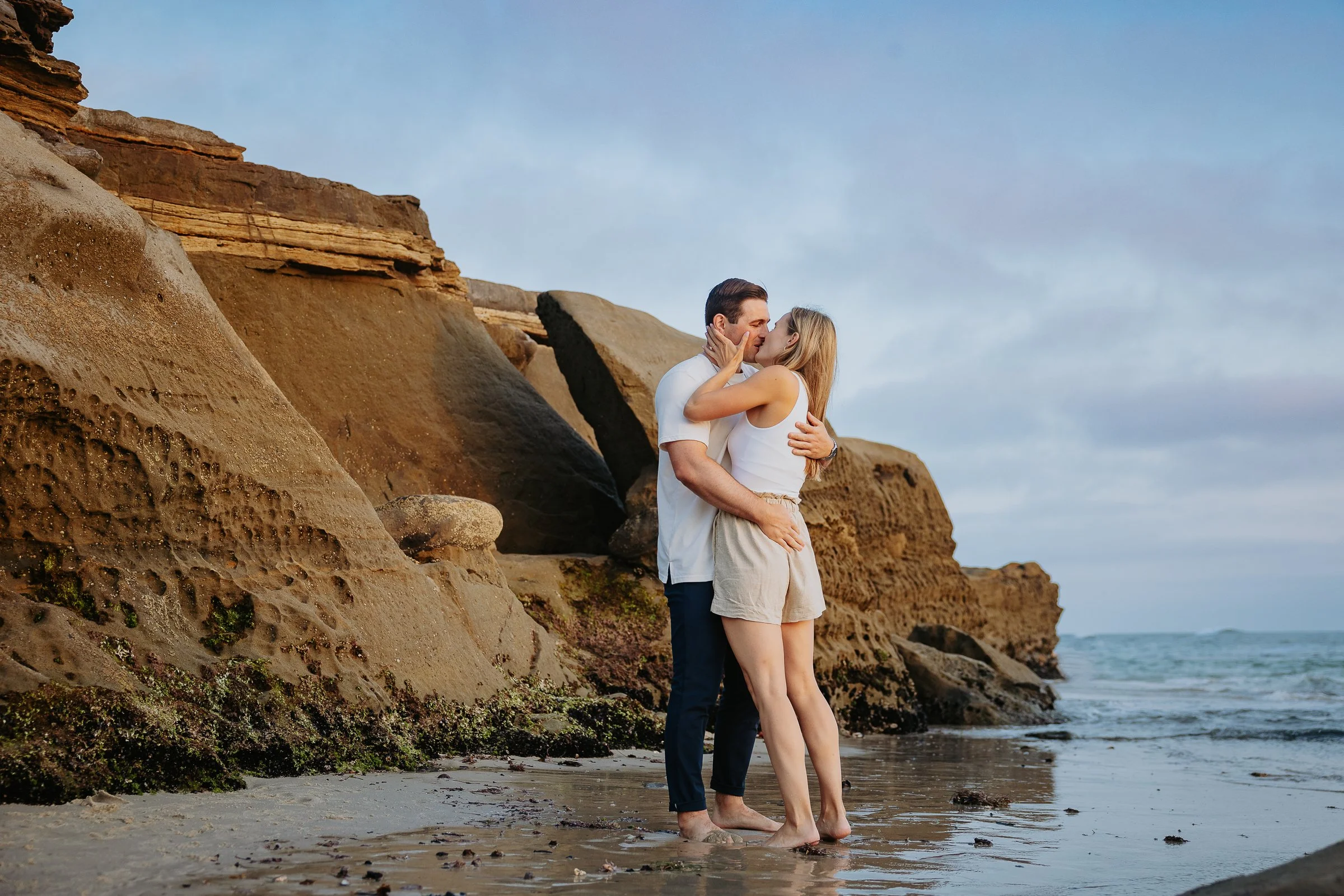 A couple hugging and kissing on the beach near rocky cliffs during sunset or sunrise.