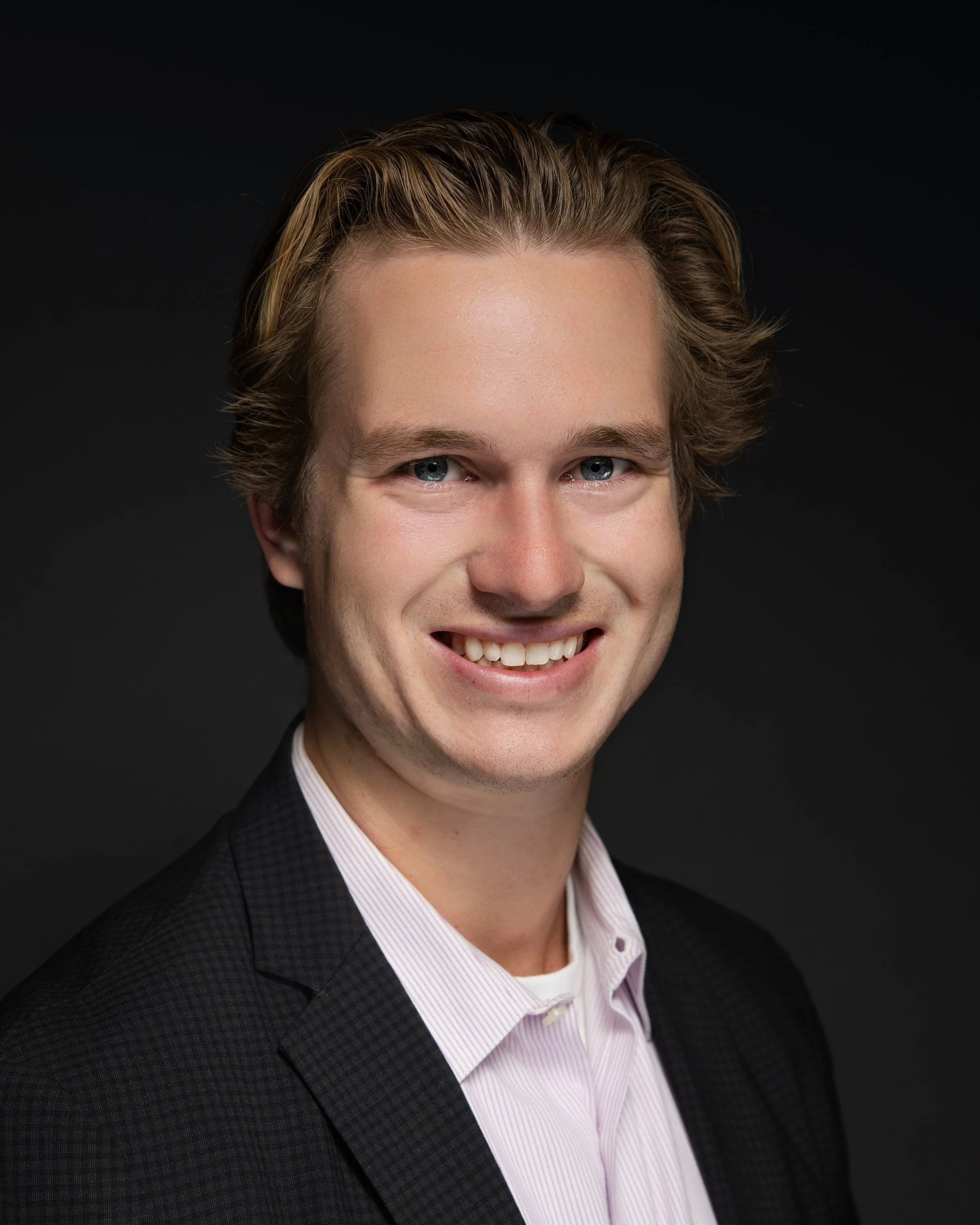 Headshot of a young man with blonde hair, blue eyes, wearing a dark blazer and a light-colored dress shirt, smiling against a dark background.