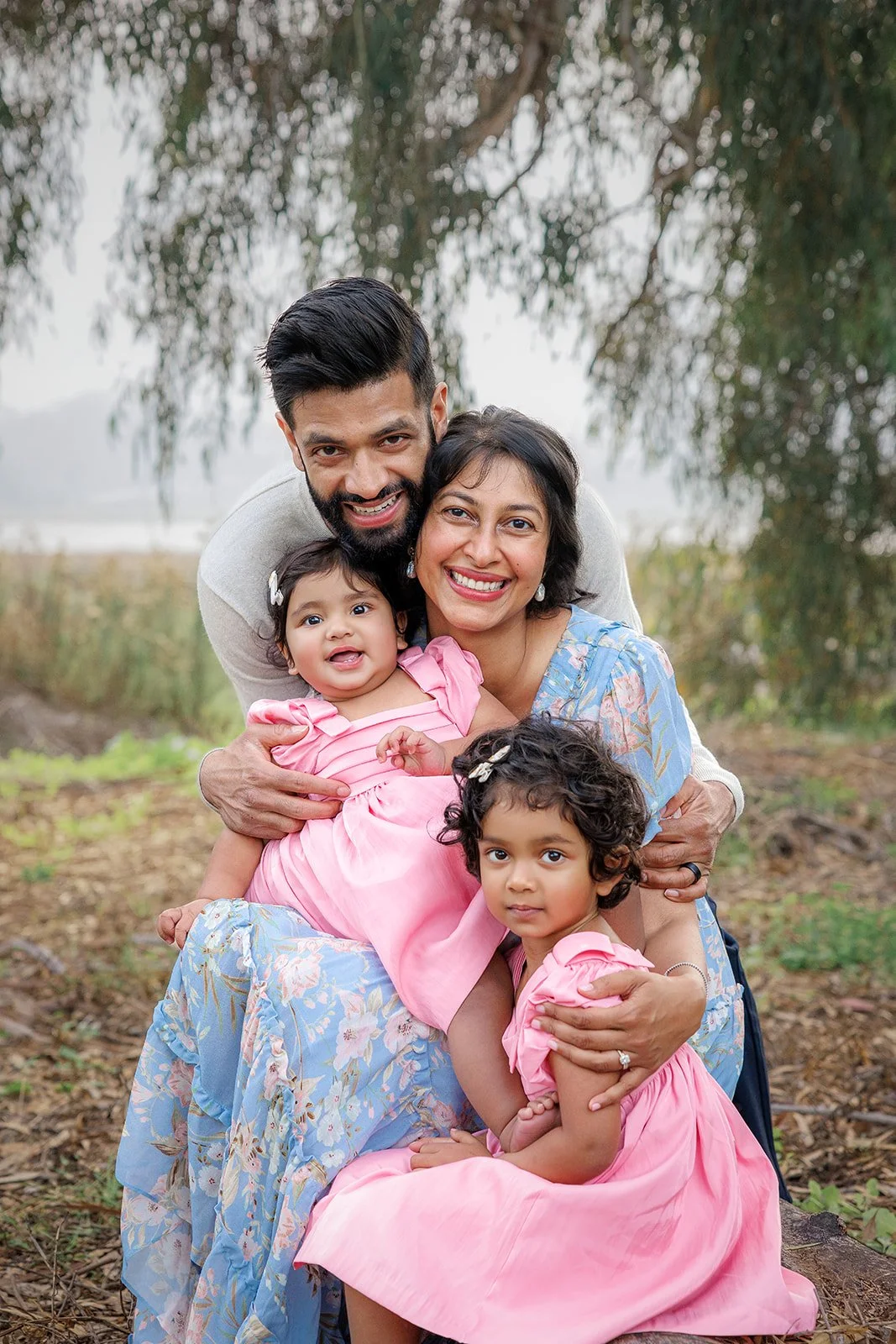A family of four, including a man, woman, and two young girls, smiling and hugging outdoors in a natural setting with trees and a slightly foggy background.