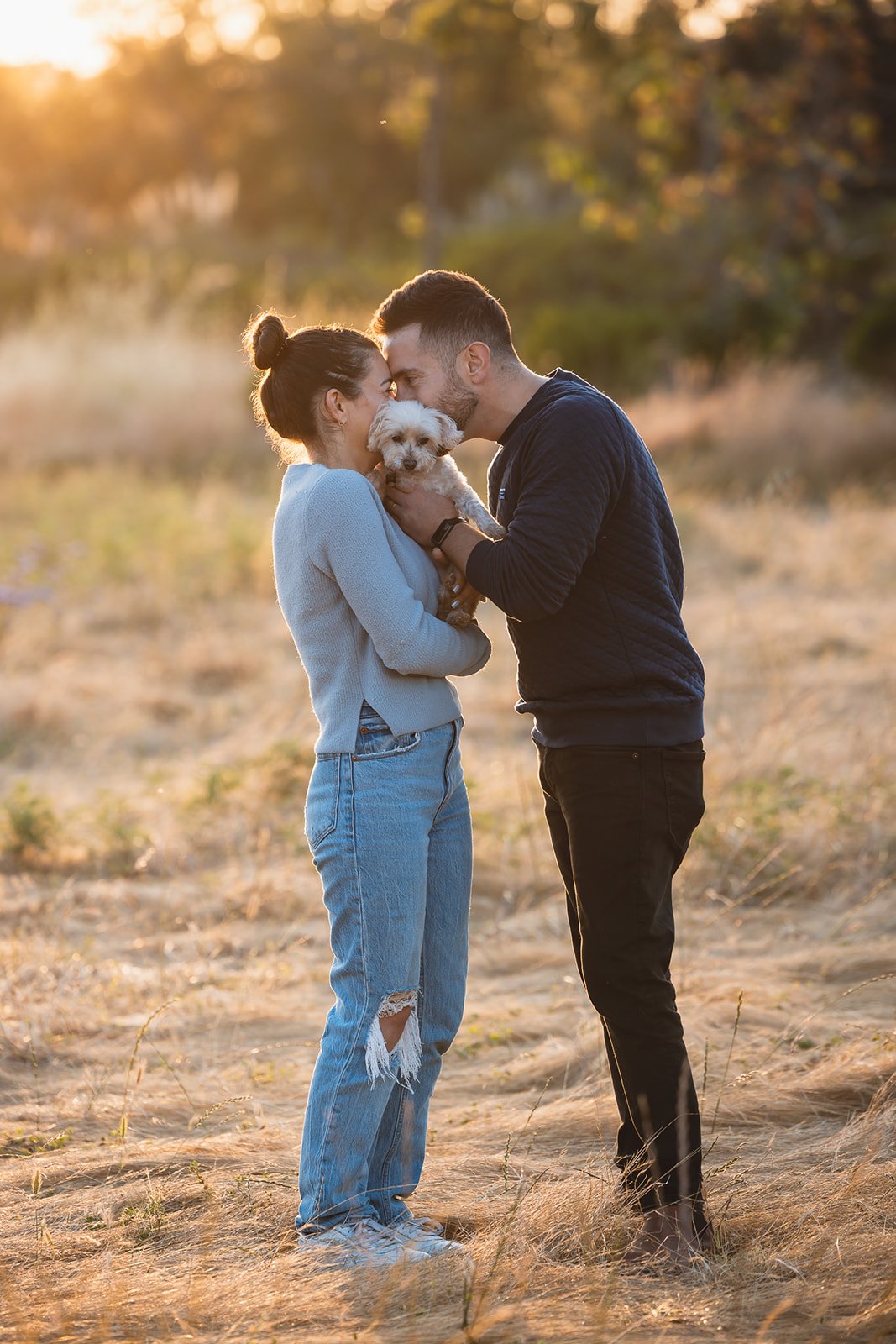 A couple standing close together outdoors at sunset, holding a small dog between them and touching foreheads affectionately.