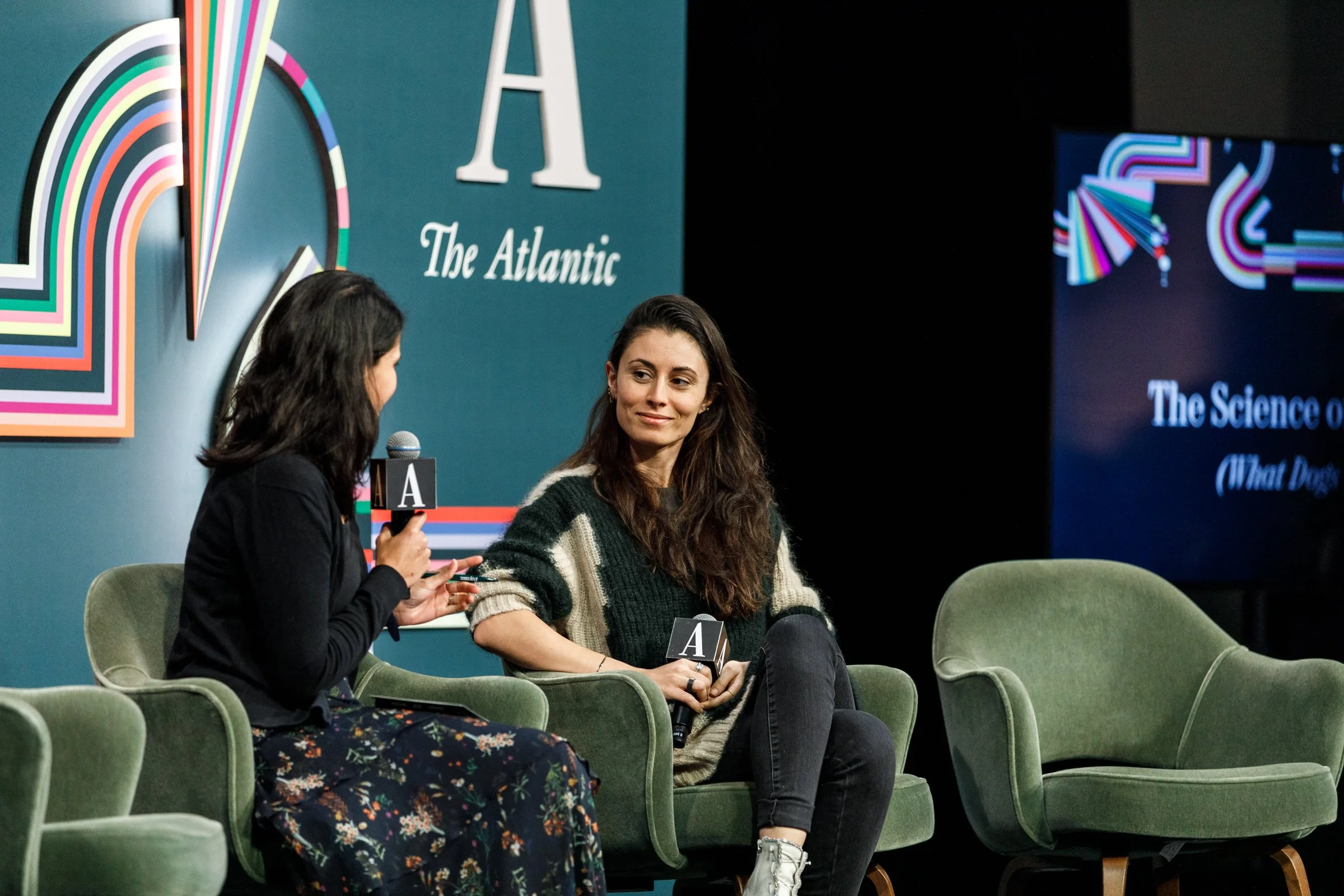 Two women seated on a stage during a discussion, one speaking into a microphone and the other listening. The stage has a blue backdrop with large text that reads 'The Atlantic' and a decorative logo. There is a large screen displaying the title 'The 